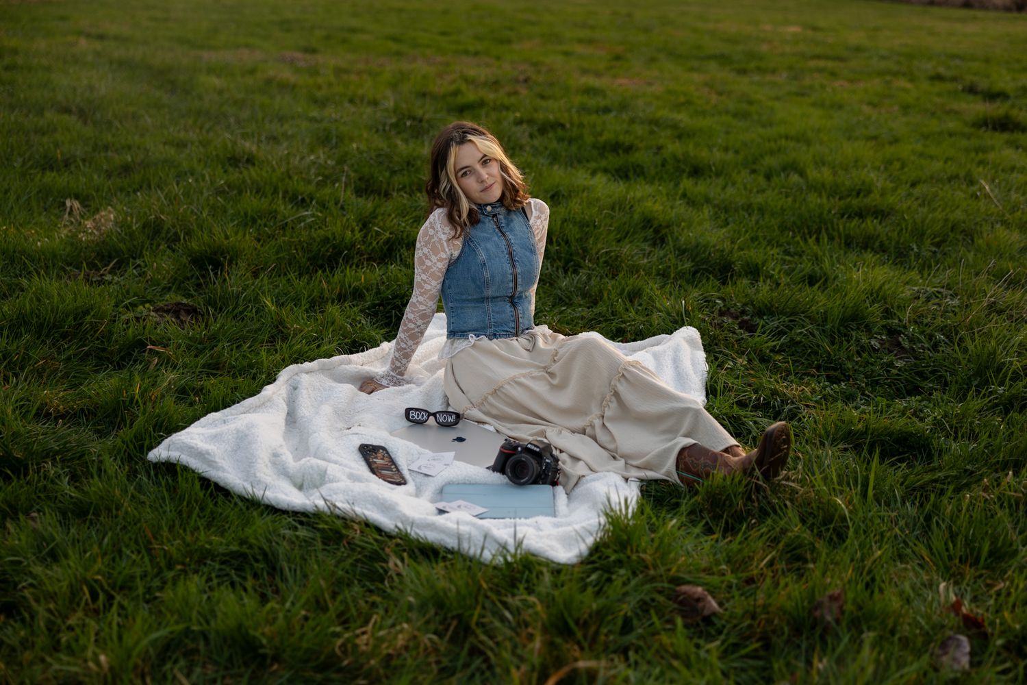 Young woman in denim jacket and cream skirt sitting on white blanket in green grass field with camera and sunglasses.