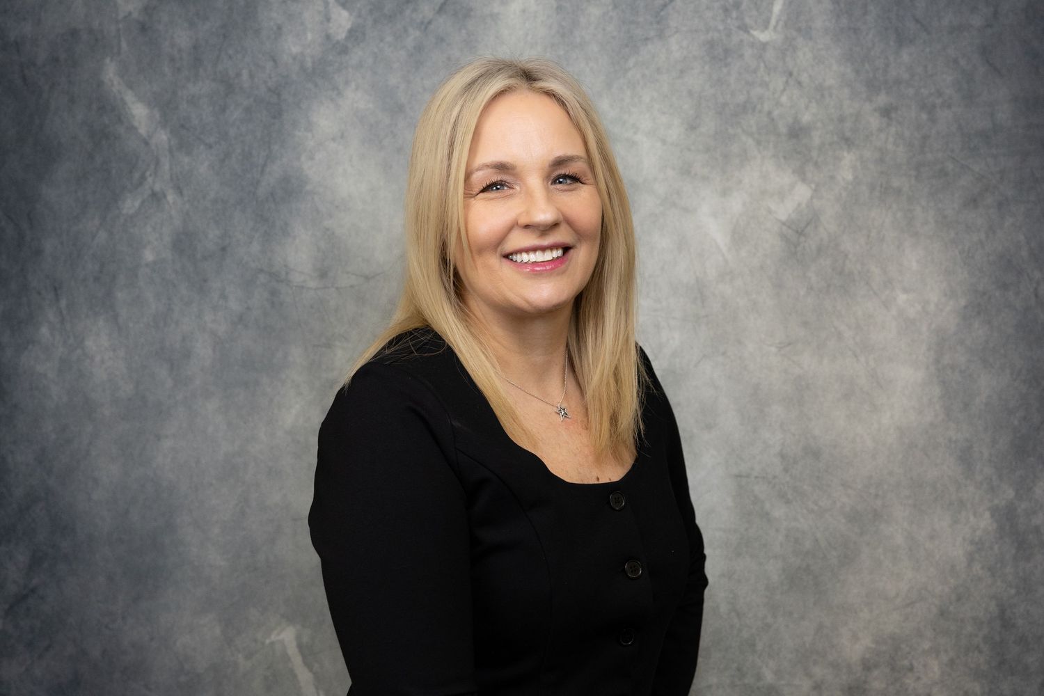Professional headshot of a smiling blonde woman in a black top against a gray studio background.