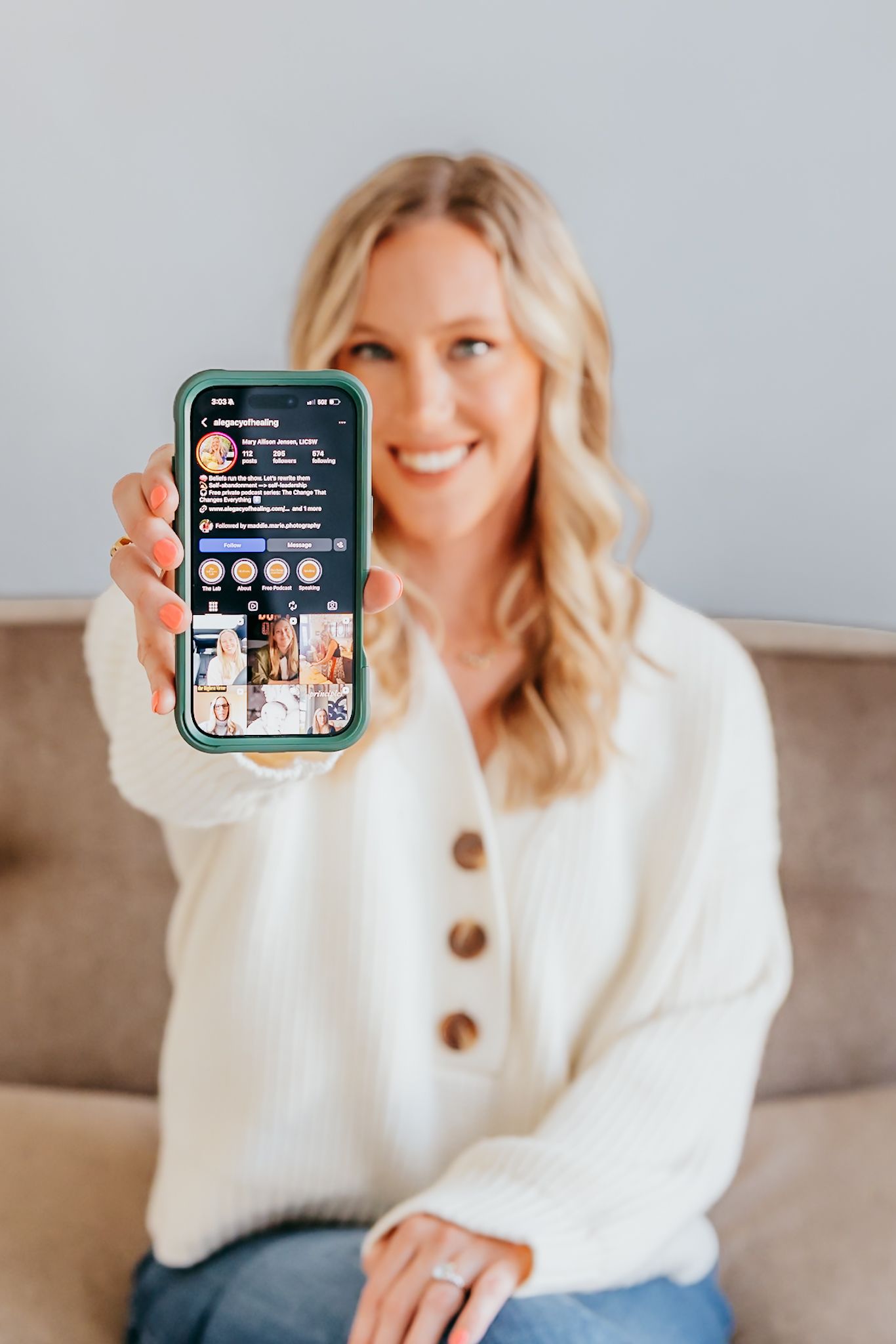 Smiling blonde woman in white sweater holding up a smartphone displaying an Instagram profile page.