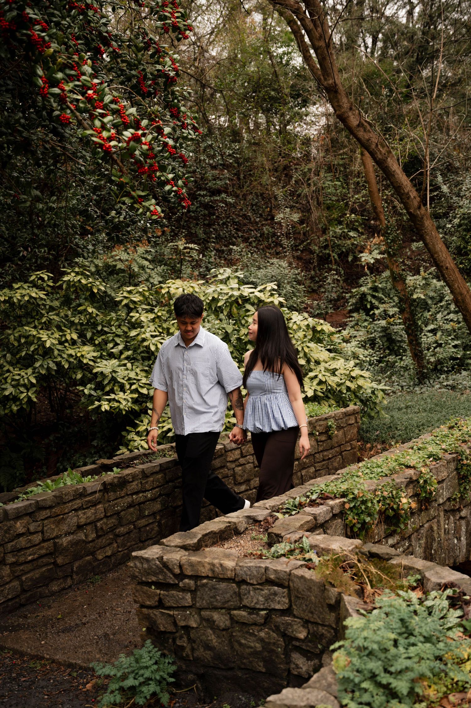 Couple walking together along a rustic stone wall path surrounded by lush greenery and flowering plants in a garden setting.