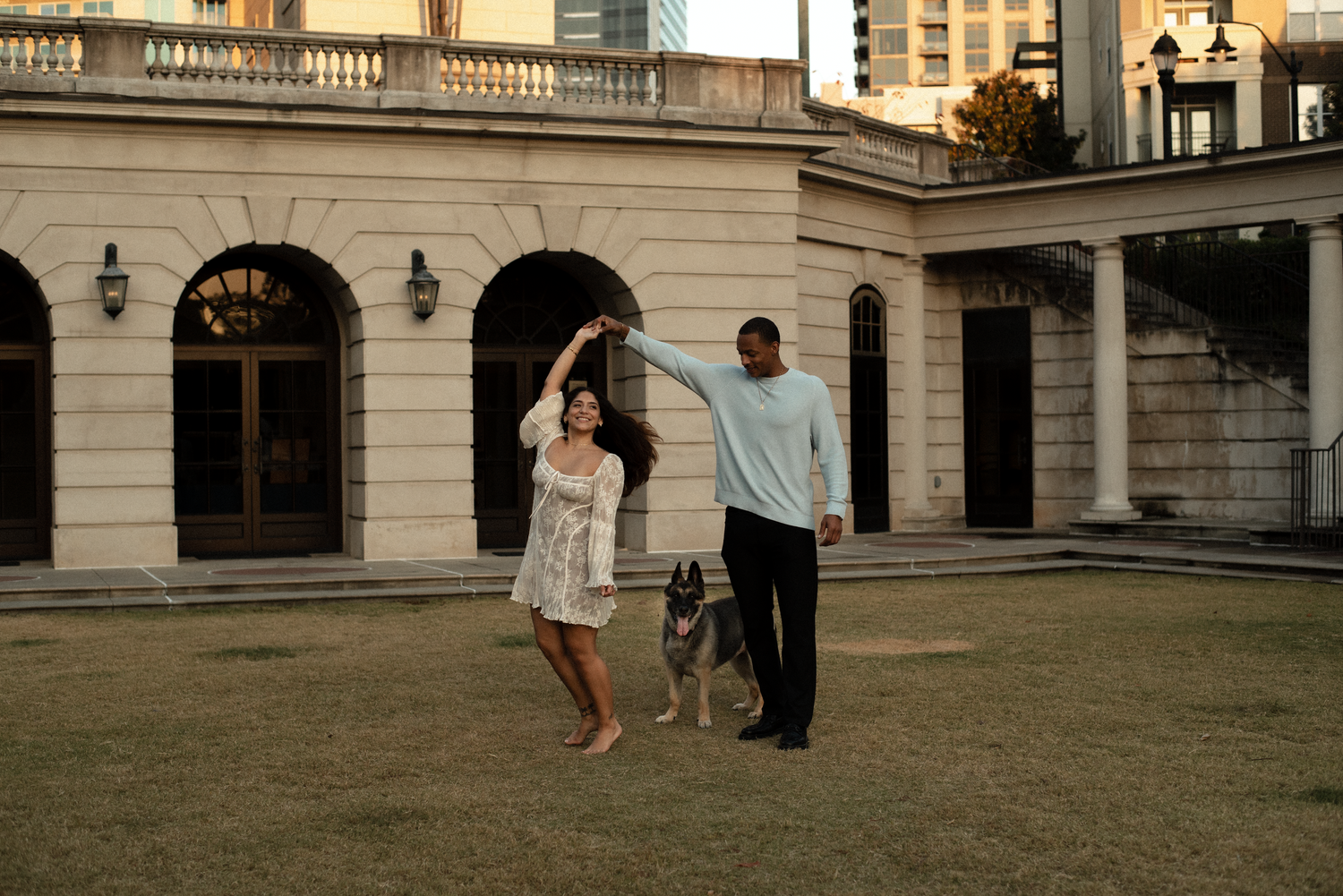 A couple walks hand in hand on a grassy lawn in front of an elegant stone building with classical architecture at sunset.