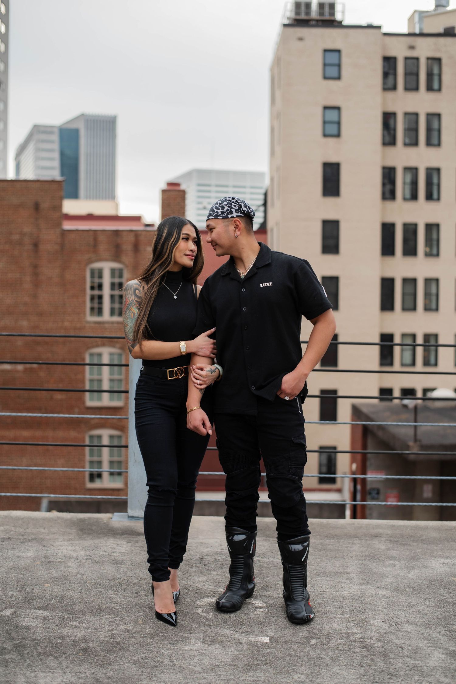 A couple in matching black outfits shares an intimate moment on an urban rooftop with city buildings in the background.