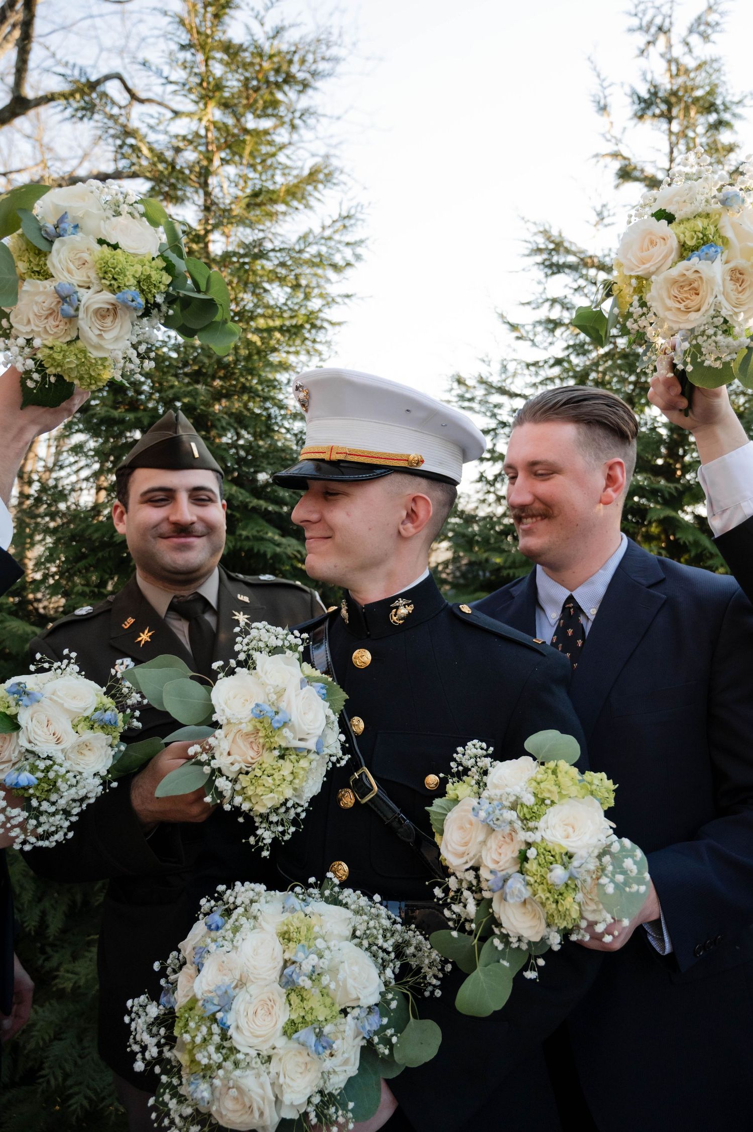 Groomsmen in formal attire hold white floral bouquets while sharing a laugh together at an outdoor wedding.