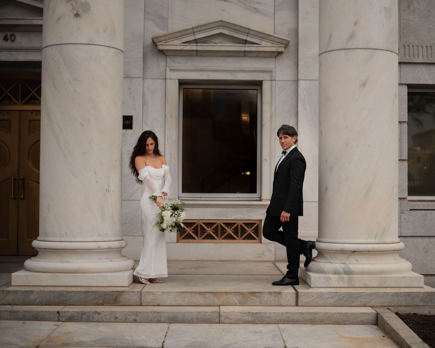 A series of architectural columns frame a romantic moment between newlyweds outside a grand building.