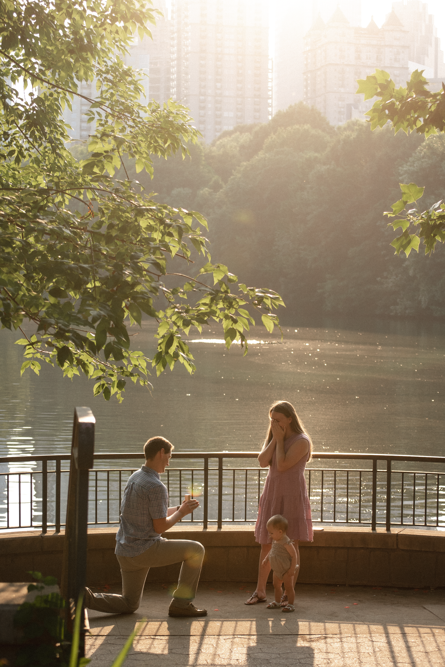 People relax on benches along a lakeside path during golden hour as sunlight filters through trees.
