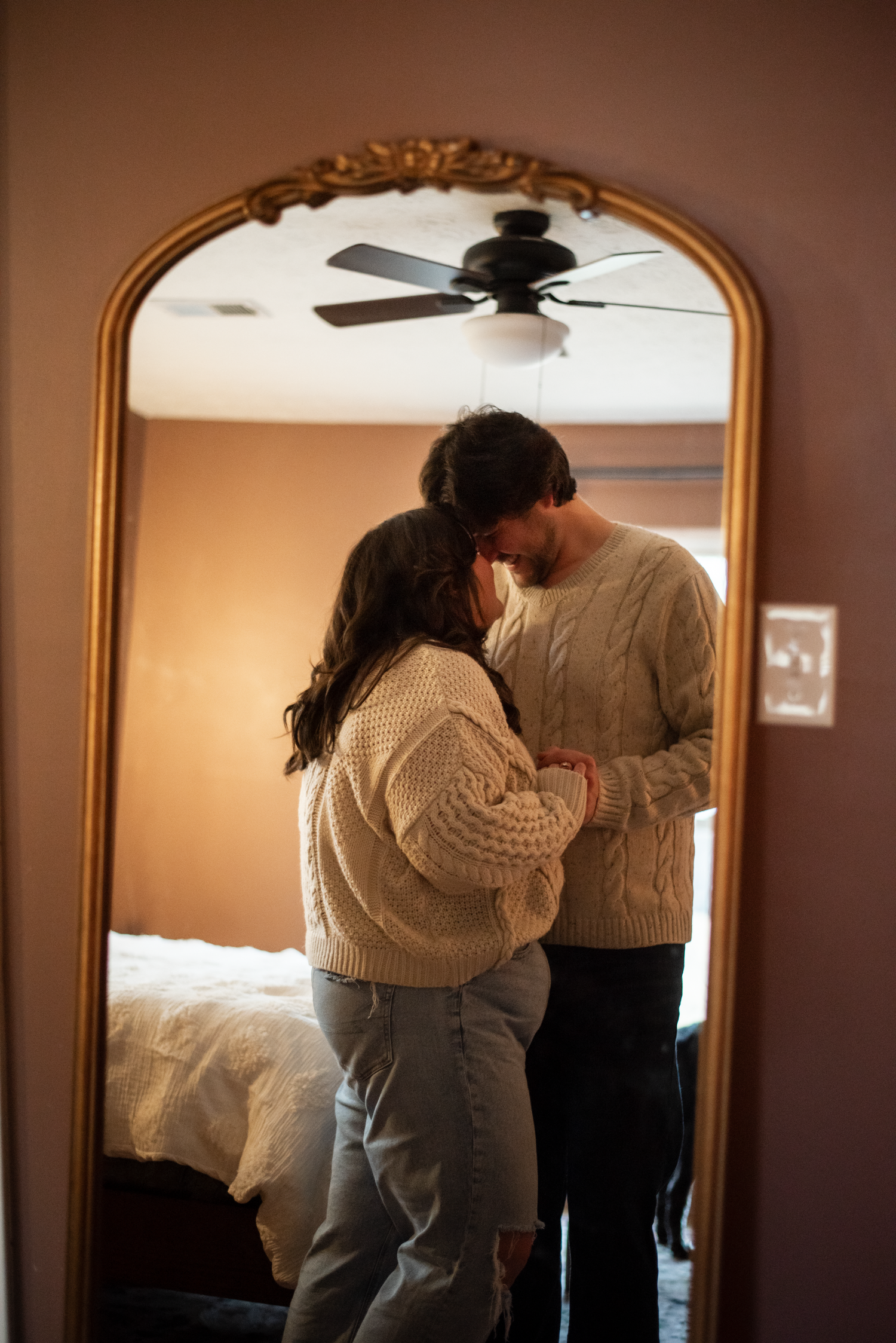 A couple in a dimly lit room share an intimate embrace in front of a large mirror.