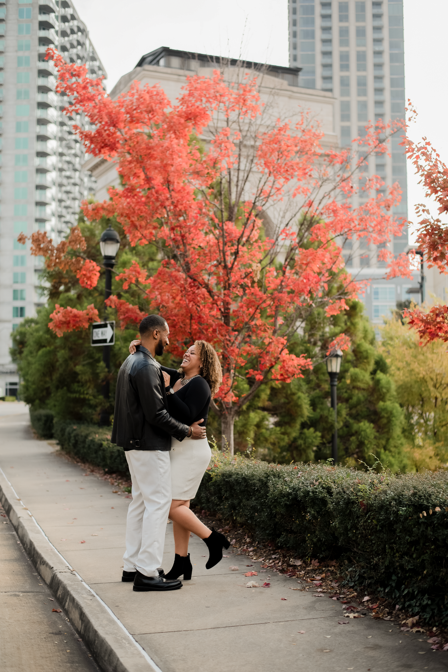 A couple embraces under vibrant red autumn trees with urban high-rise buildings in the background.