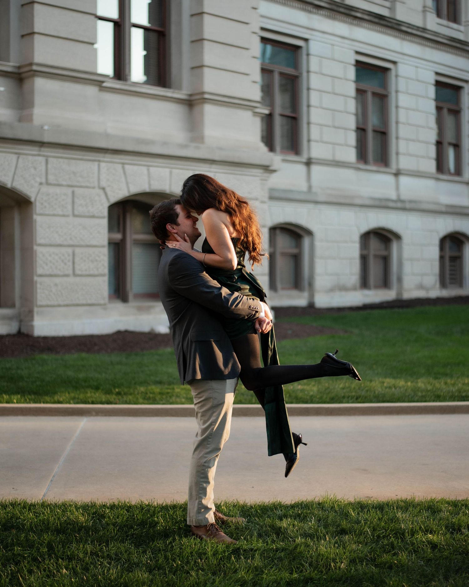 Romantic couple embracing on a grassy lawn outside a grand stone building.