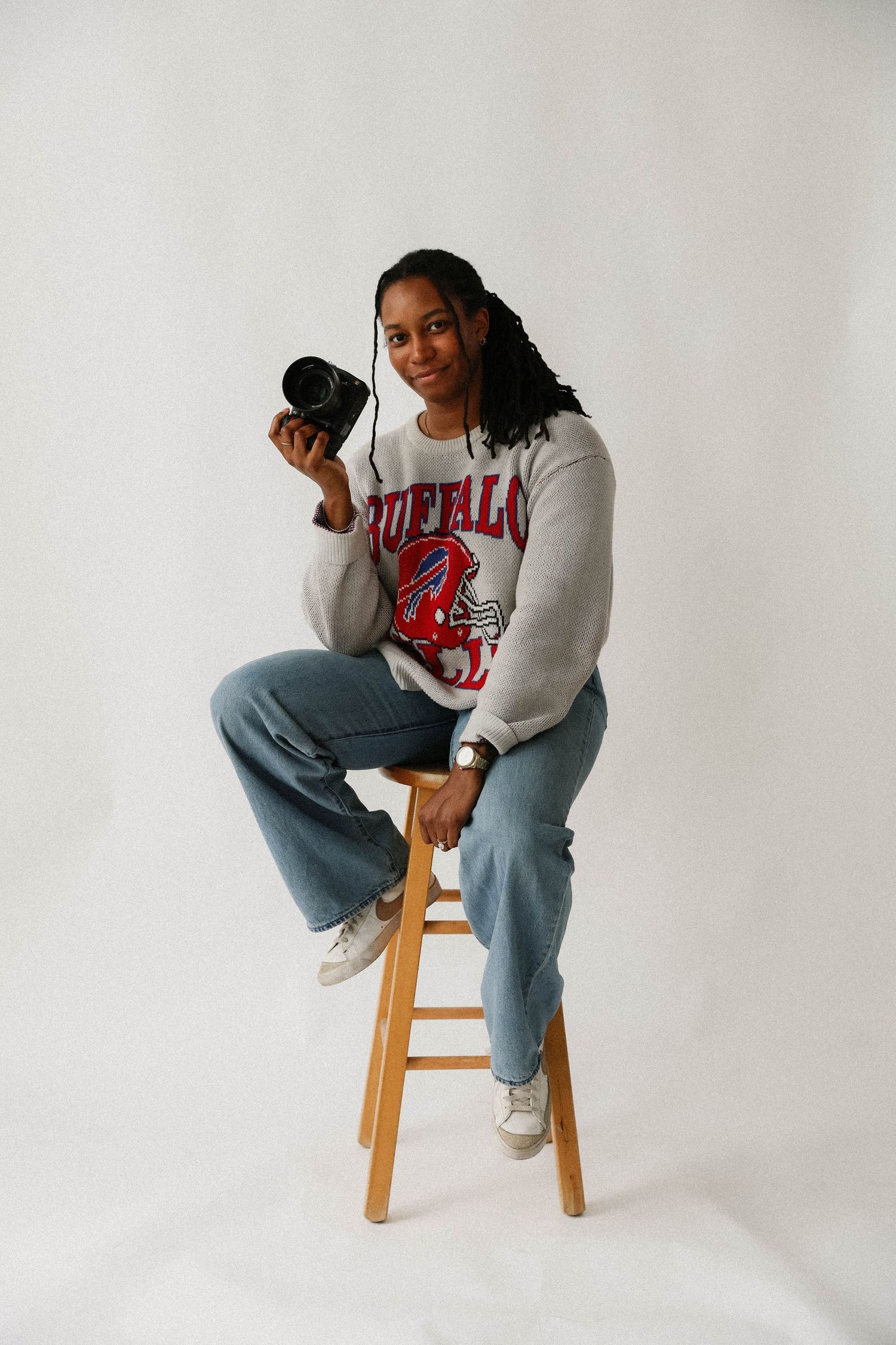 A person in a Chicago Bulls sweatshirt and jeans sits casually on a wooden stool against a white backdrop.