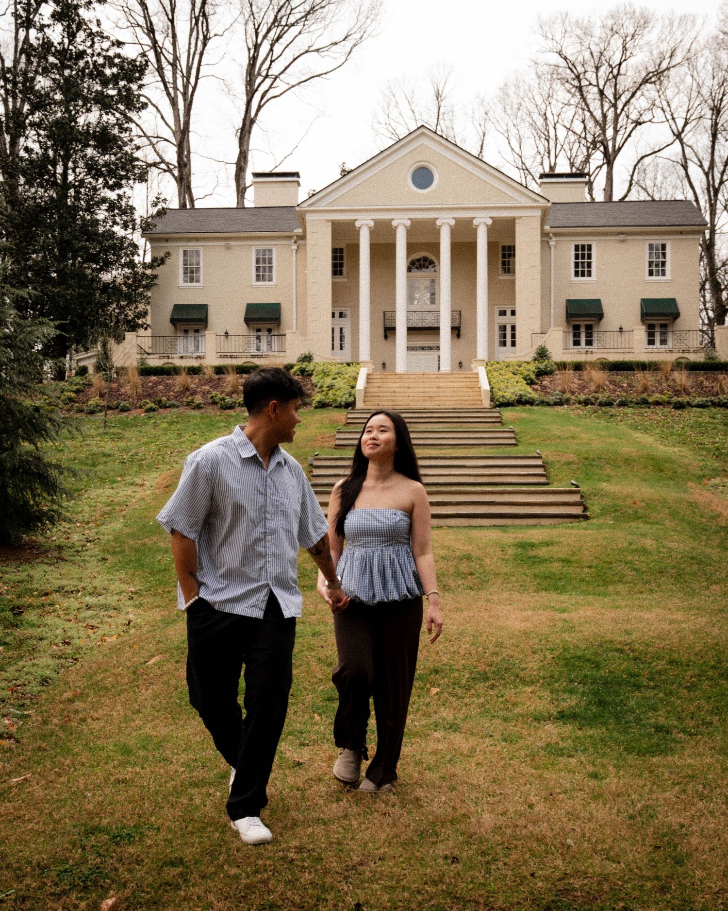 A couple walks together on the lawn in front of a grand Southern colonial mansion with white columns and symmetrical design.