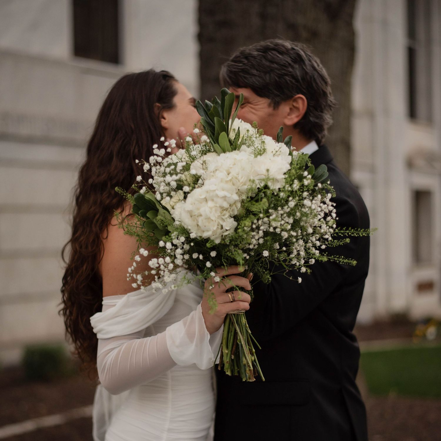 A couple in wedding attire shares an intimate moment while holding a white floral bouquet.