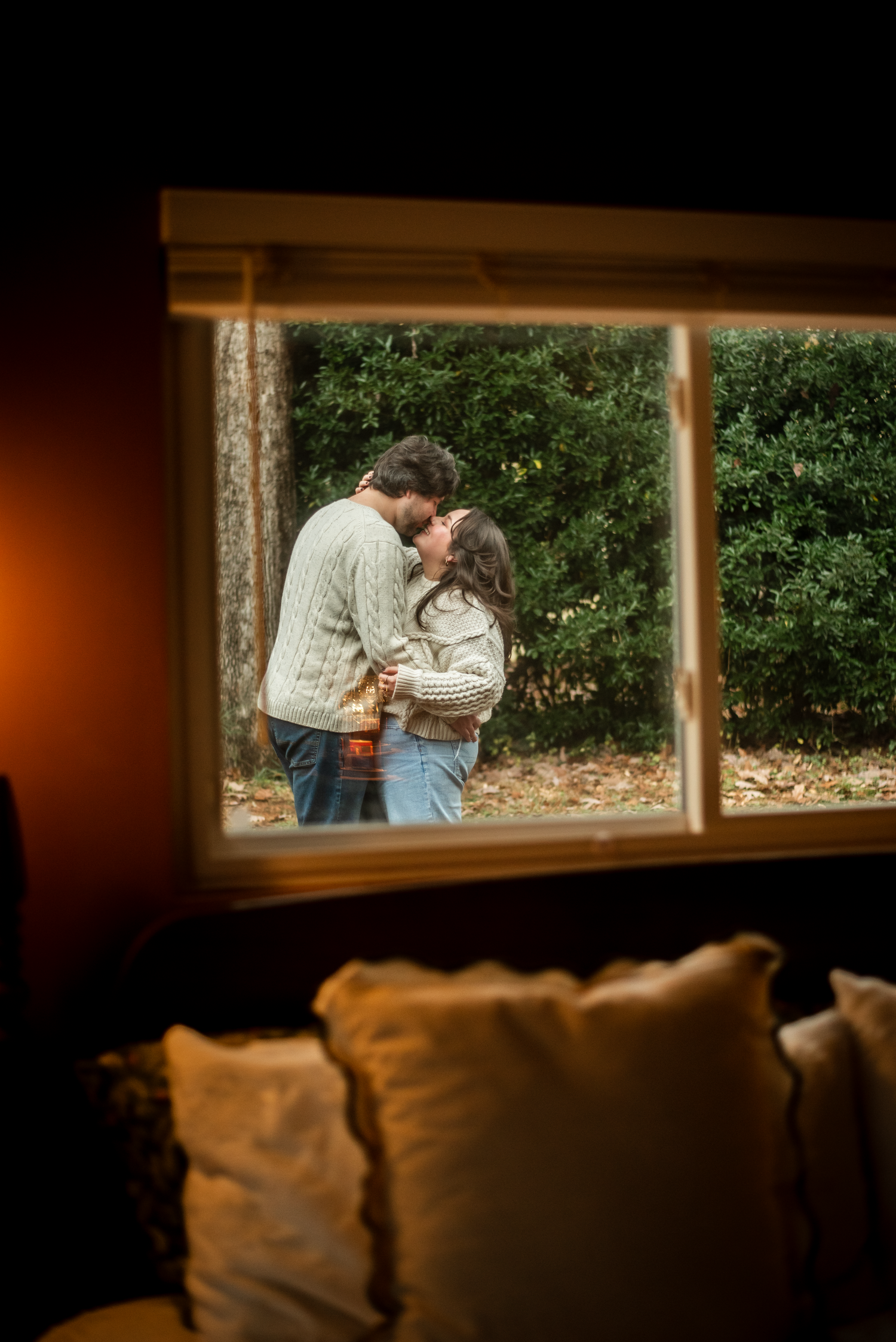 A romantic scene viewed through a panoramic window shows a couple embracing in front of lush greenery outdoors.