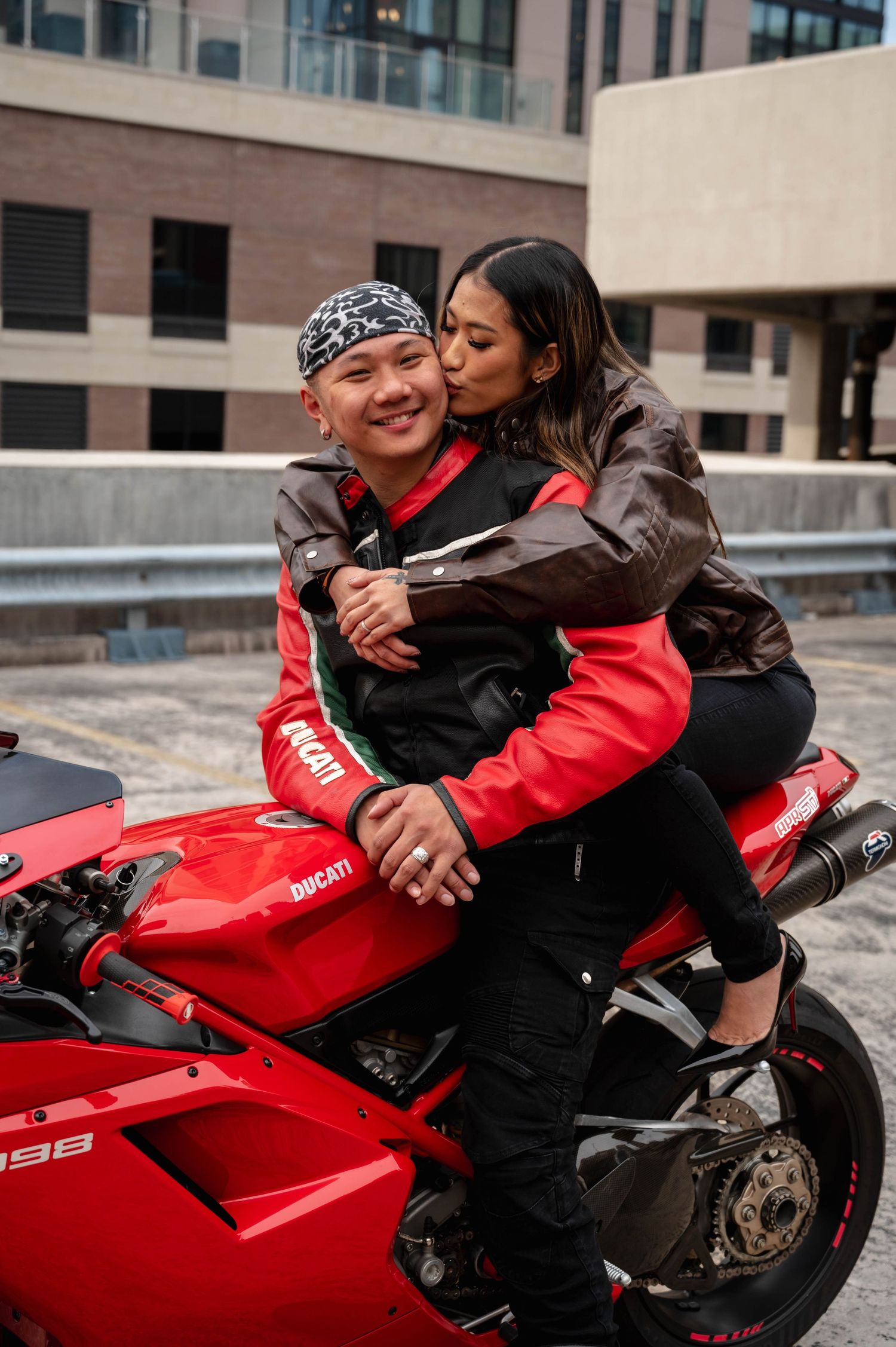 Two people share an intimate moment while sitting on a bright red motorcycle parked in an urban setting.