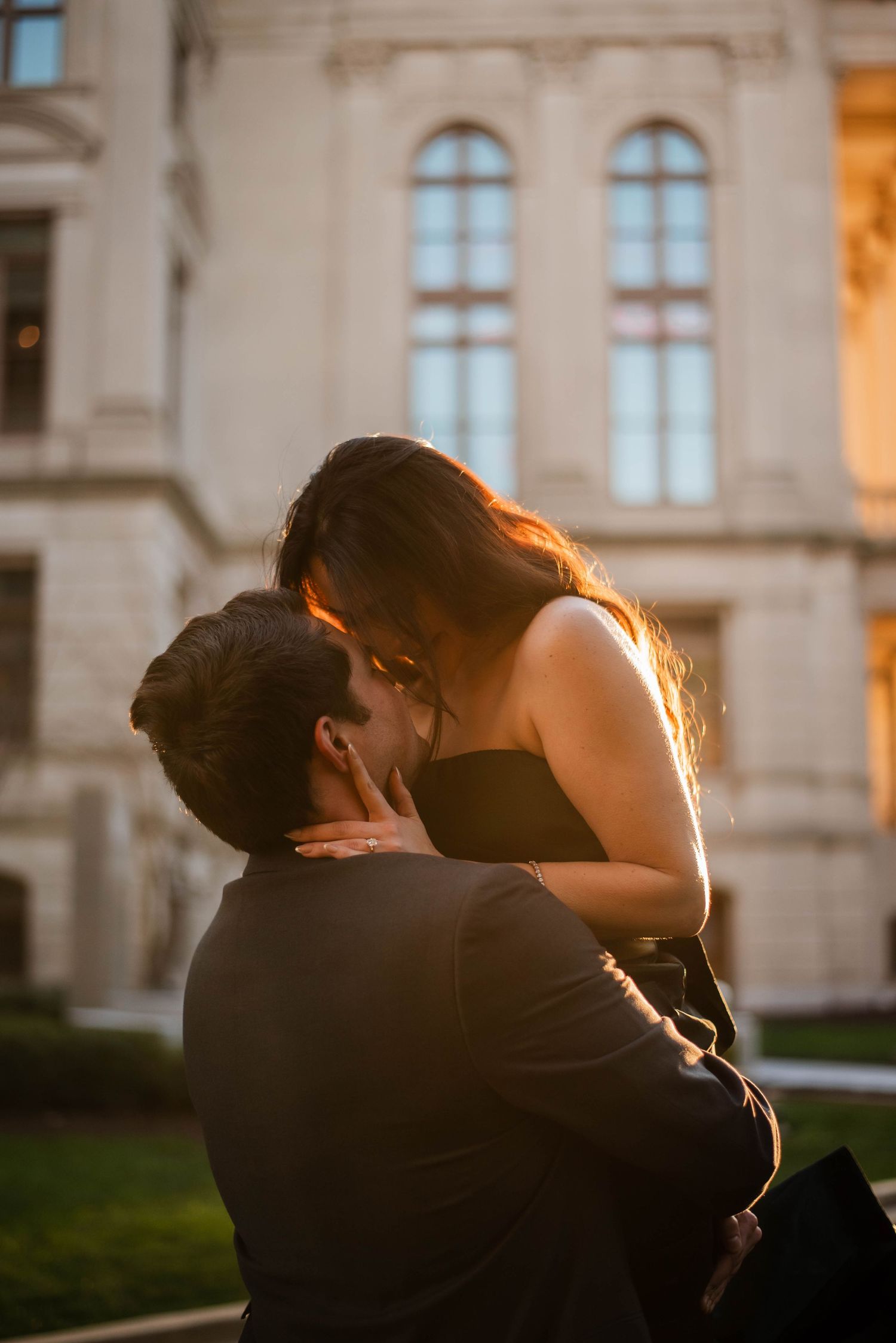A romantic couple embraces and kisses in front of a grand building during golden hour sunset.