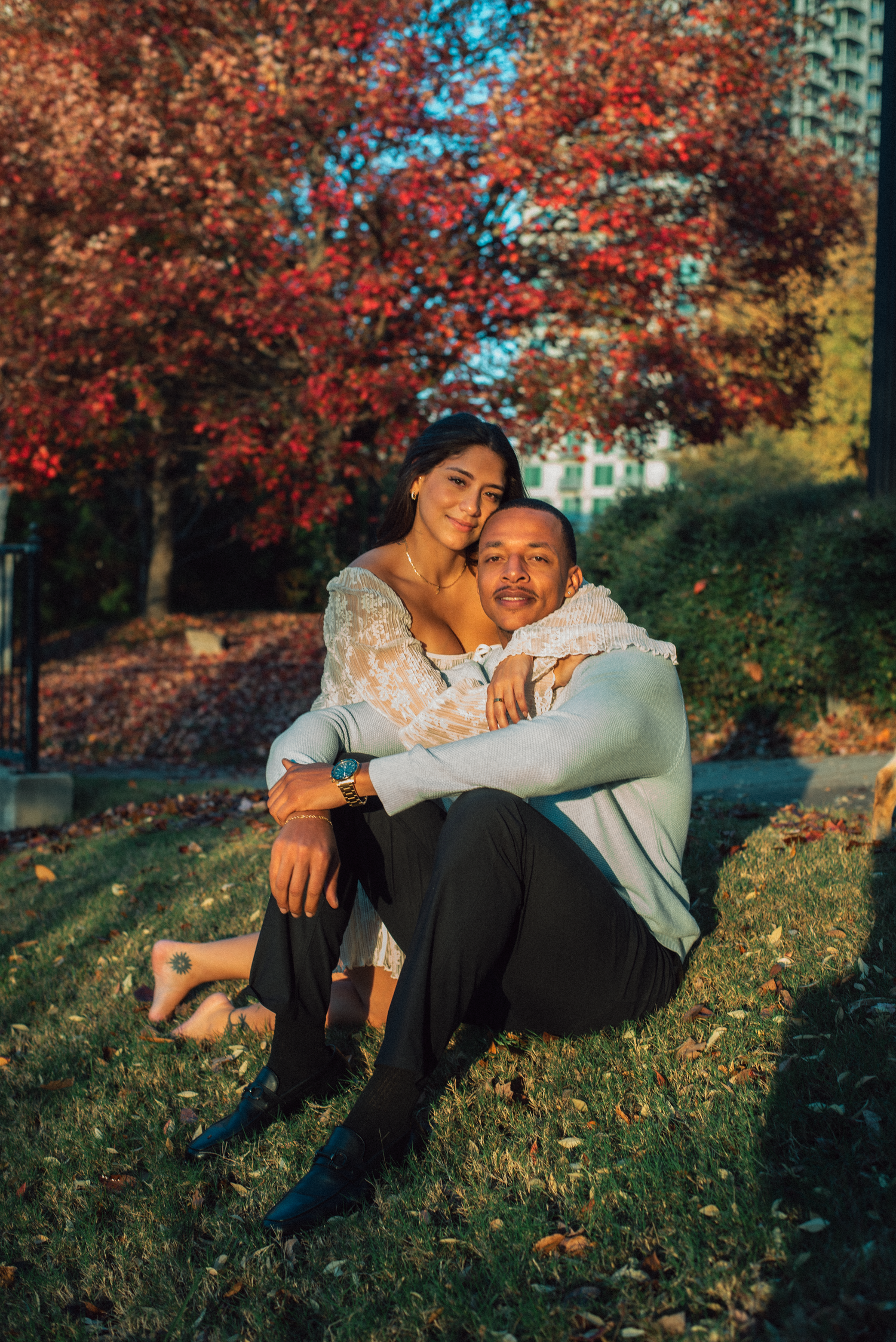 A couple sits together on grass during autumn with vibrant red maple trees and blue sky in the background.