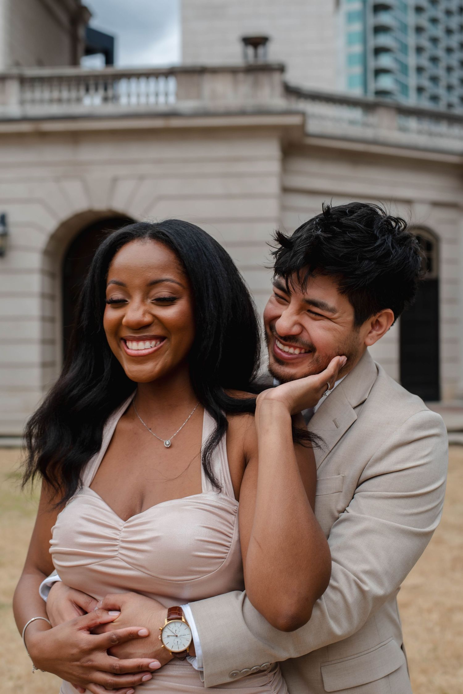 A romantic couple shares joyful laughter and embraces during an outdoor photoshoot in front of a grand building.