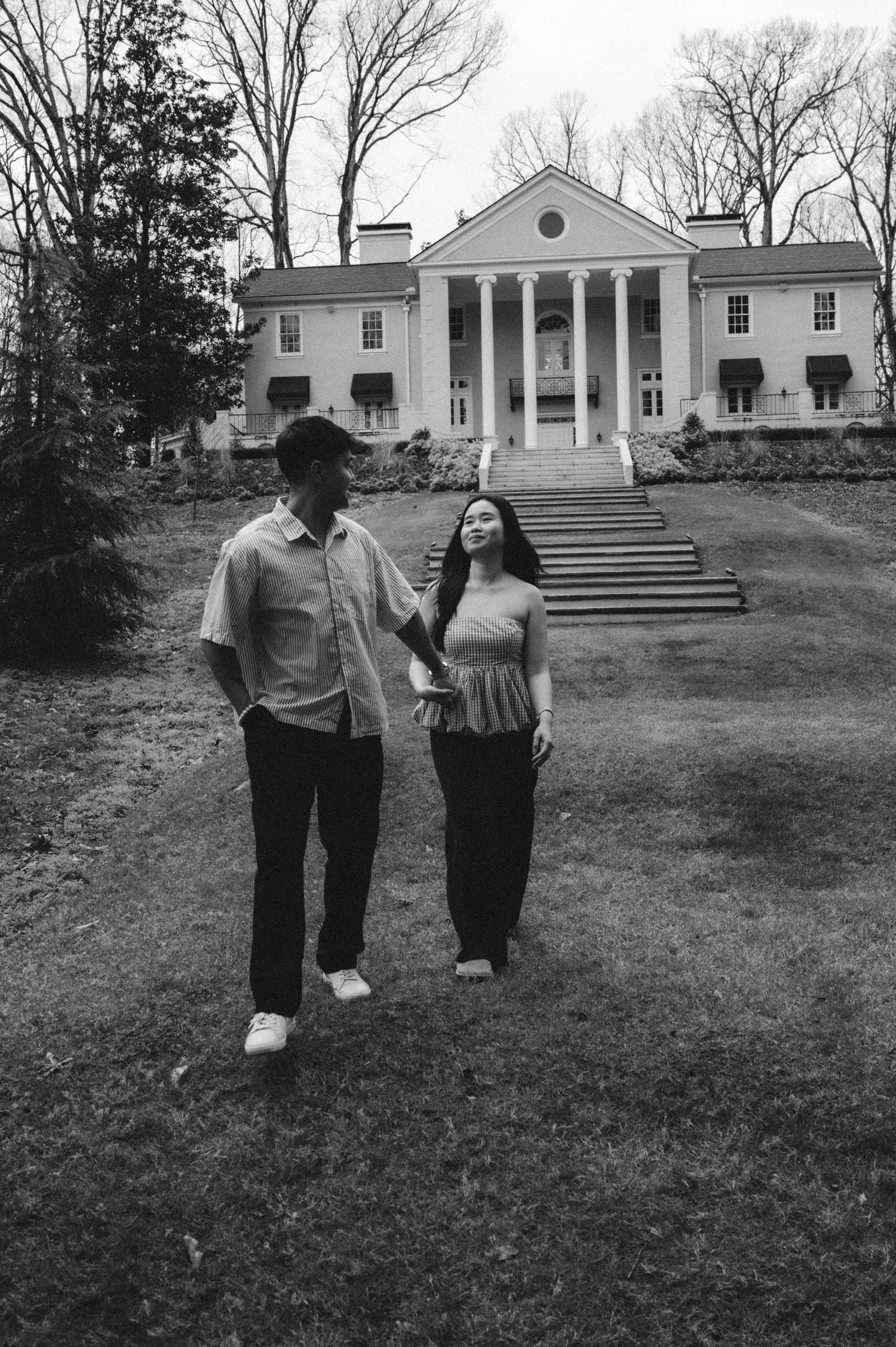 A black and white photograph showing a couple standing on a lawn in front of a stately Southern colonial-style mansion.