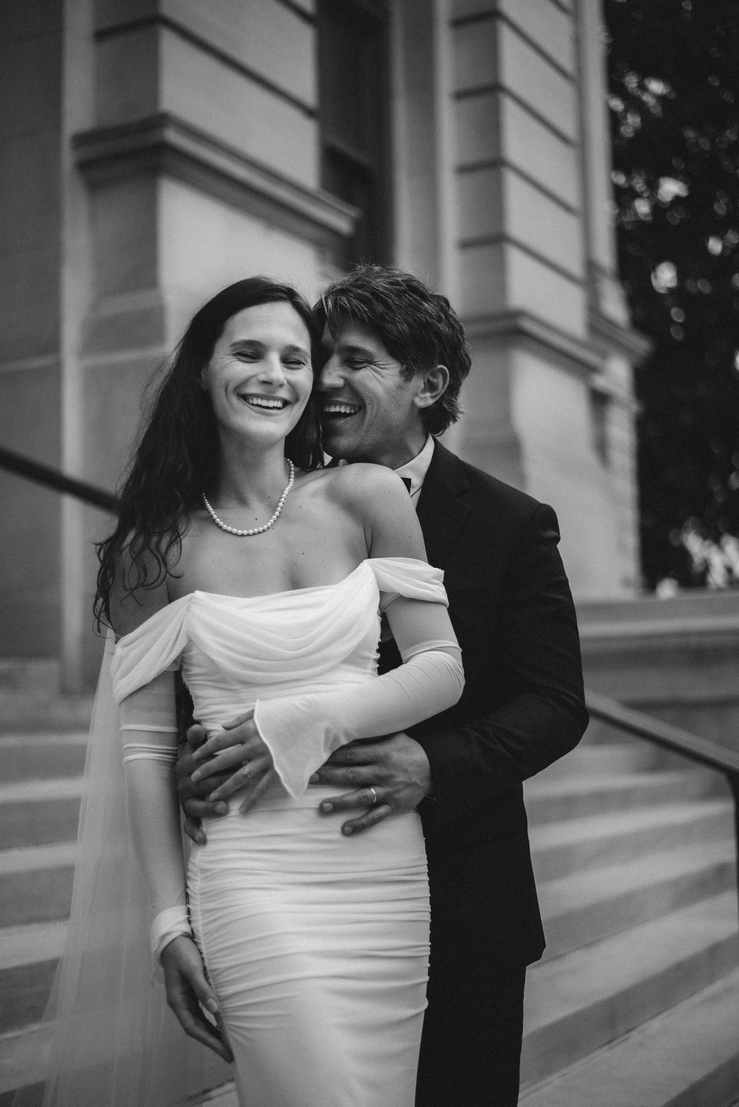 A couple in formal wedding attire embraces on elegant stone steps in a romantic black and white photograph.