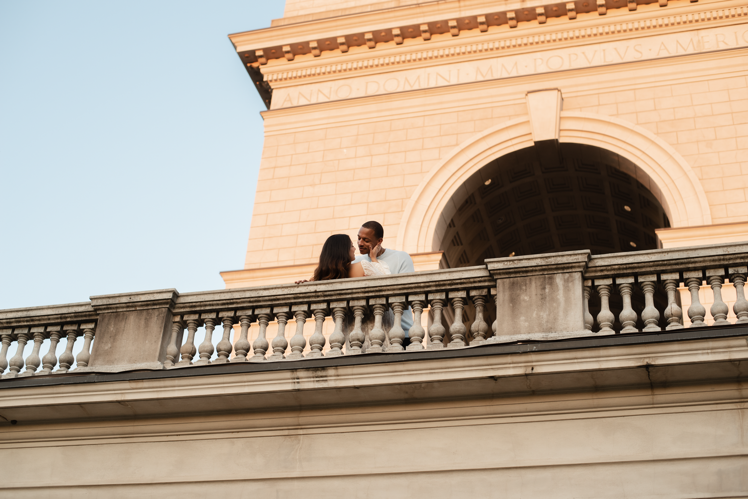 Two silhouettes stand together on an elegant balcony with arched architecture during golden hour.
