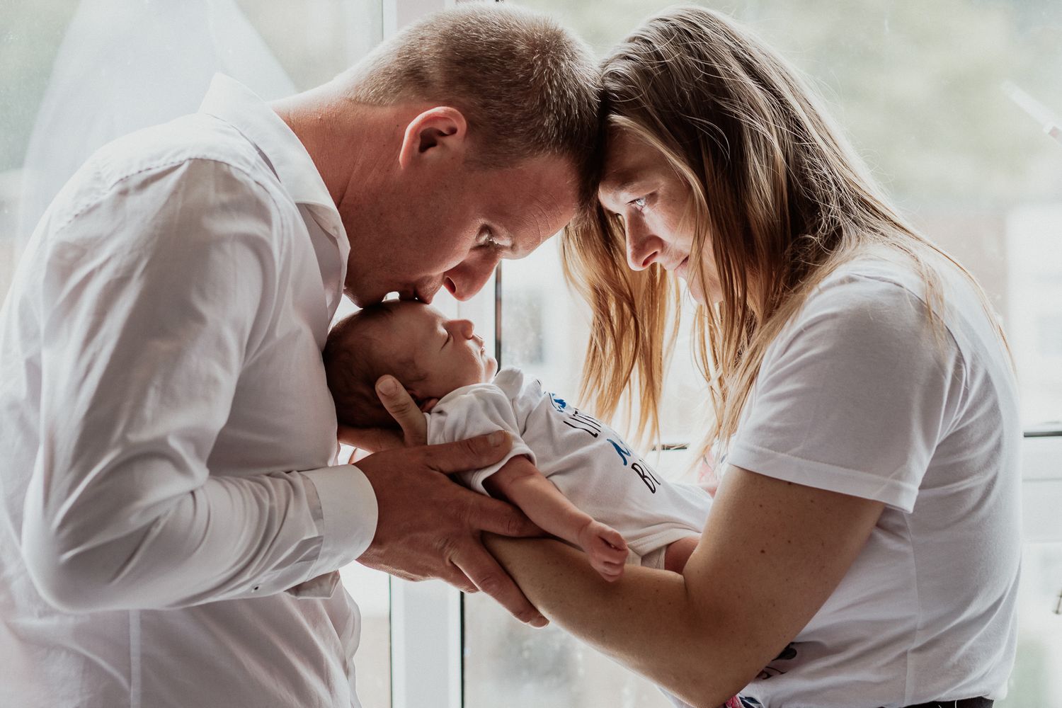 Emotional photo of mother, father and new born child. Father kissing forehead of baby during new born session in family home in Kent