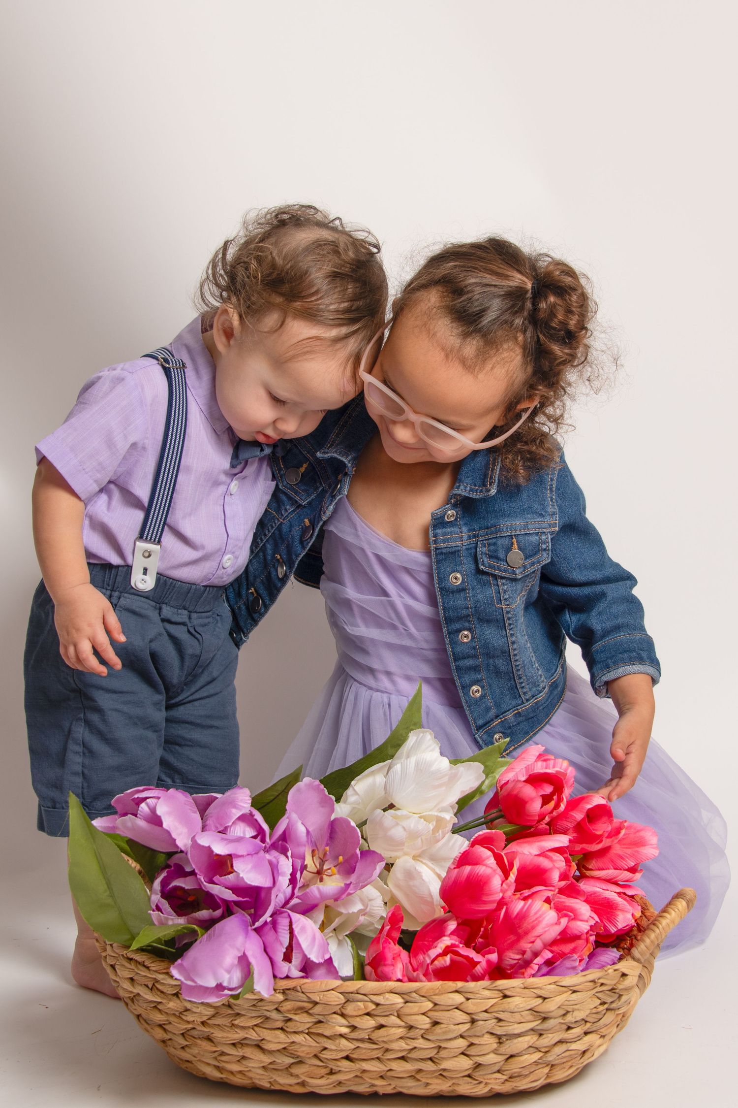 Two toddlers in matching outfits lean over a woven basket filled with colorful spring tulips during a photo session.
