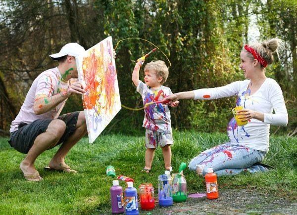 Family painting outdoors with colorful paints, toddler holding hula hoop while parents engage in creative art activity on green lawn.