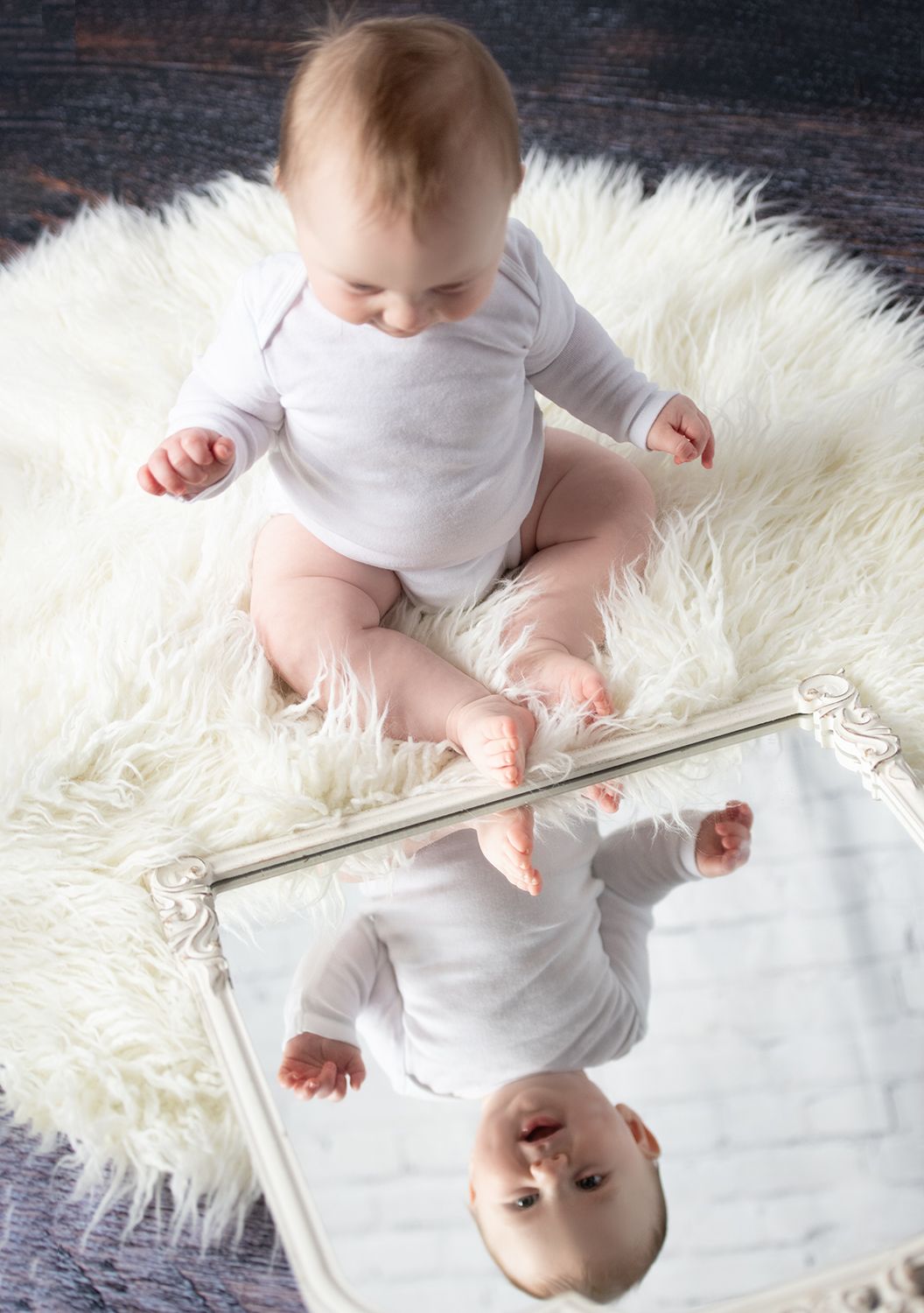 Baby in white onesie lying on soft white fur blanket while looking at his image in a mirror