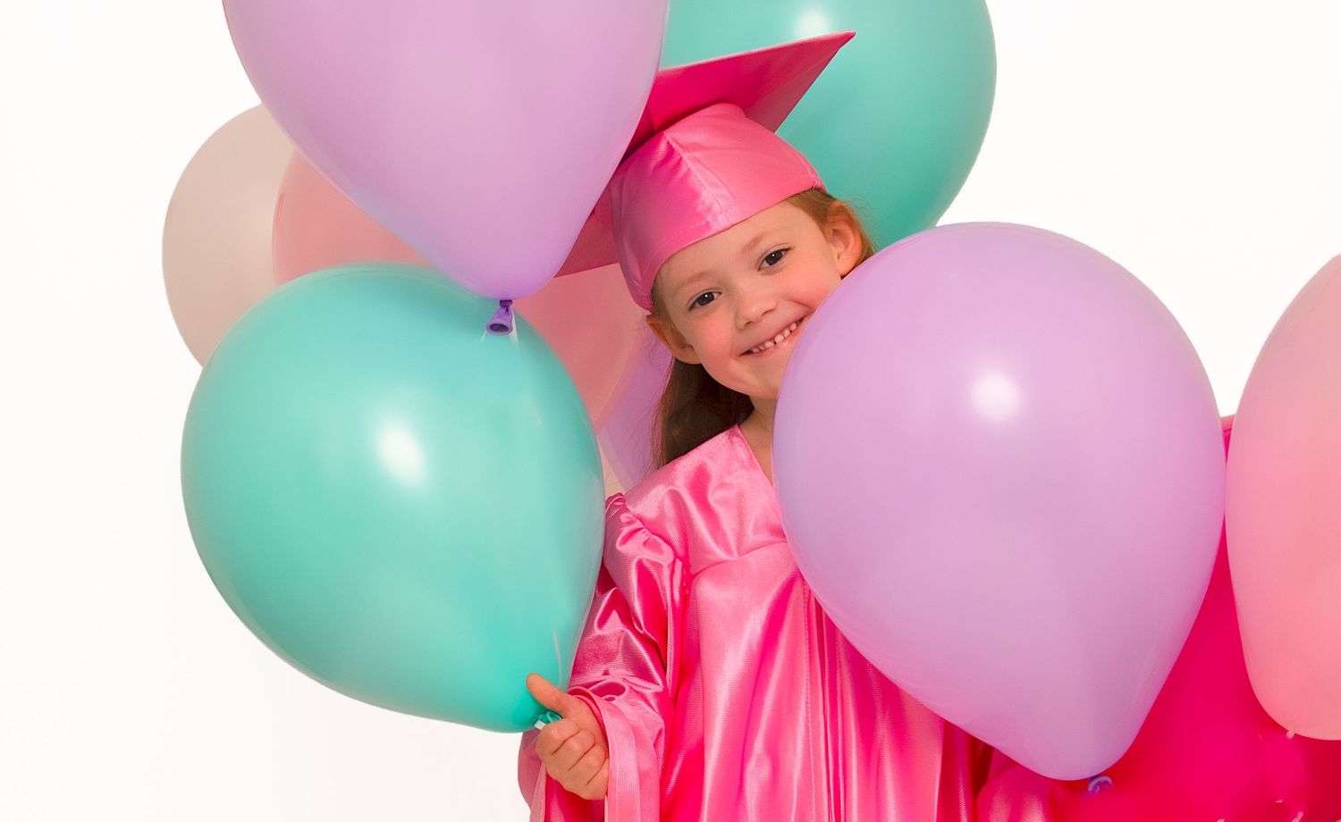 Young girl in pink graduation cap and gown smiling behind colorful balloons on white background.