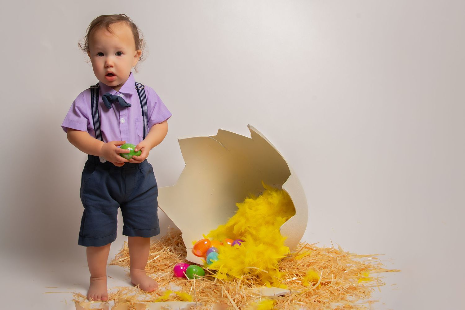 A child in purple shirt and navy shorts plays with colorful Easter decorations and yellow tissue paper on the floor.