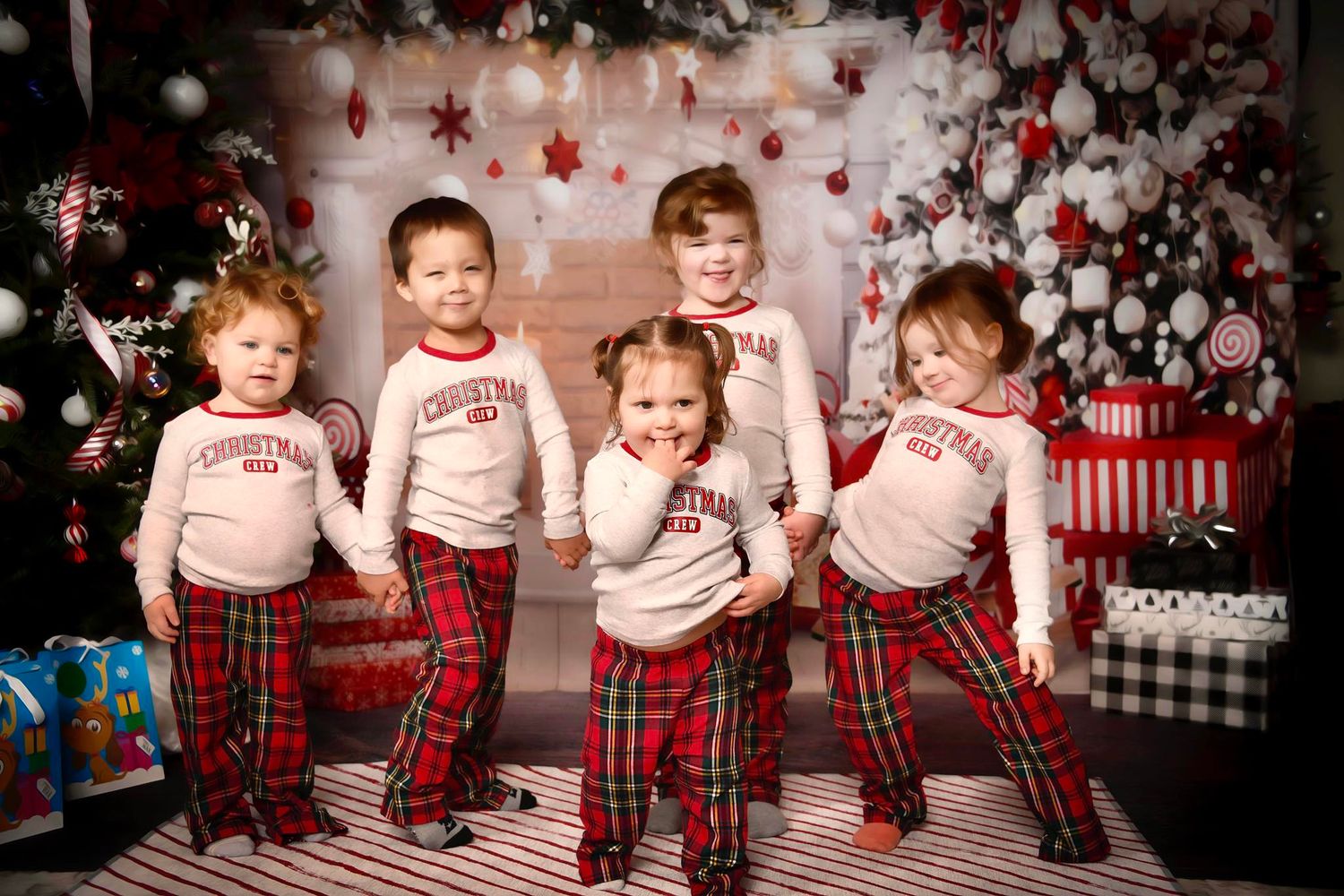 Children in matching plaid pajamas pose together in front of a festive holiday backdrop with snowflakes and ornaments.