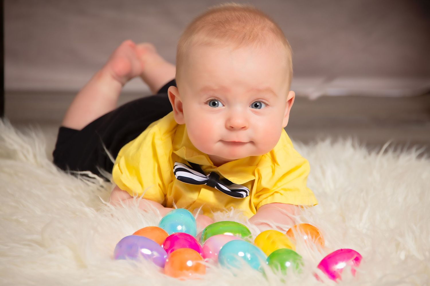 A baby lies on a white furry rug surrounded by colorful plastic Easter eggs.