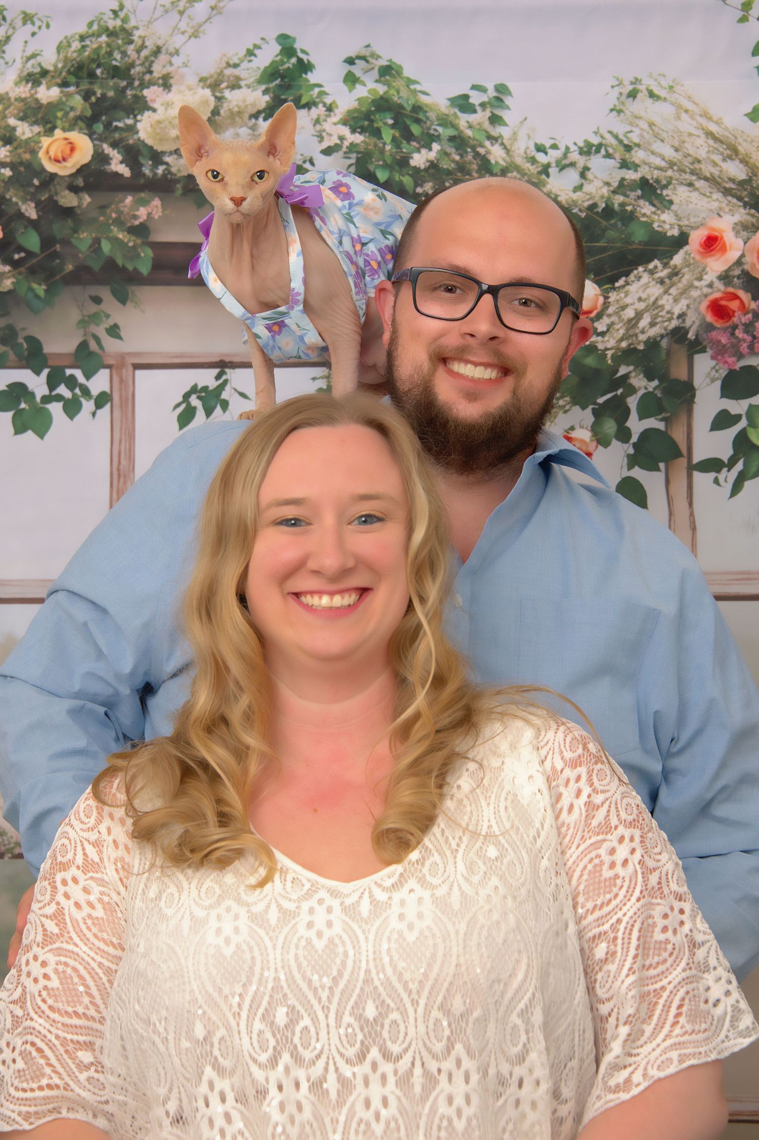 Smiling couple in blue and white outfits posing with a Sphynx cat in a dress against a floral backdrop.
