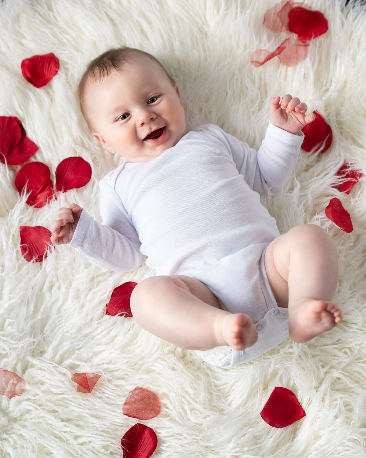 Baby in white onesie lying on fluffy white rug with scattered red rose petals creates romantic Valentine's Day scene.
