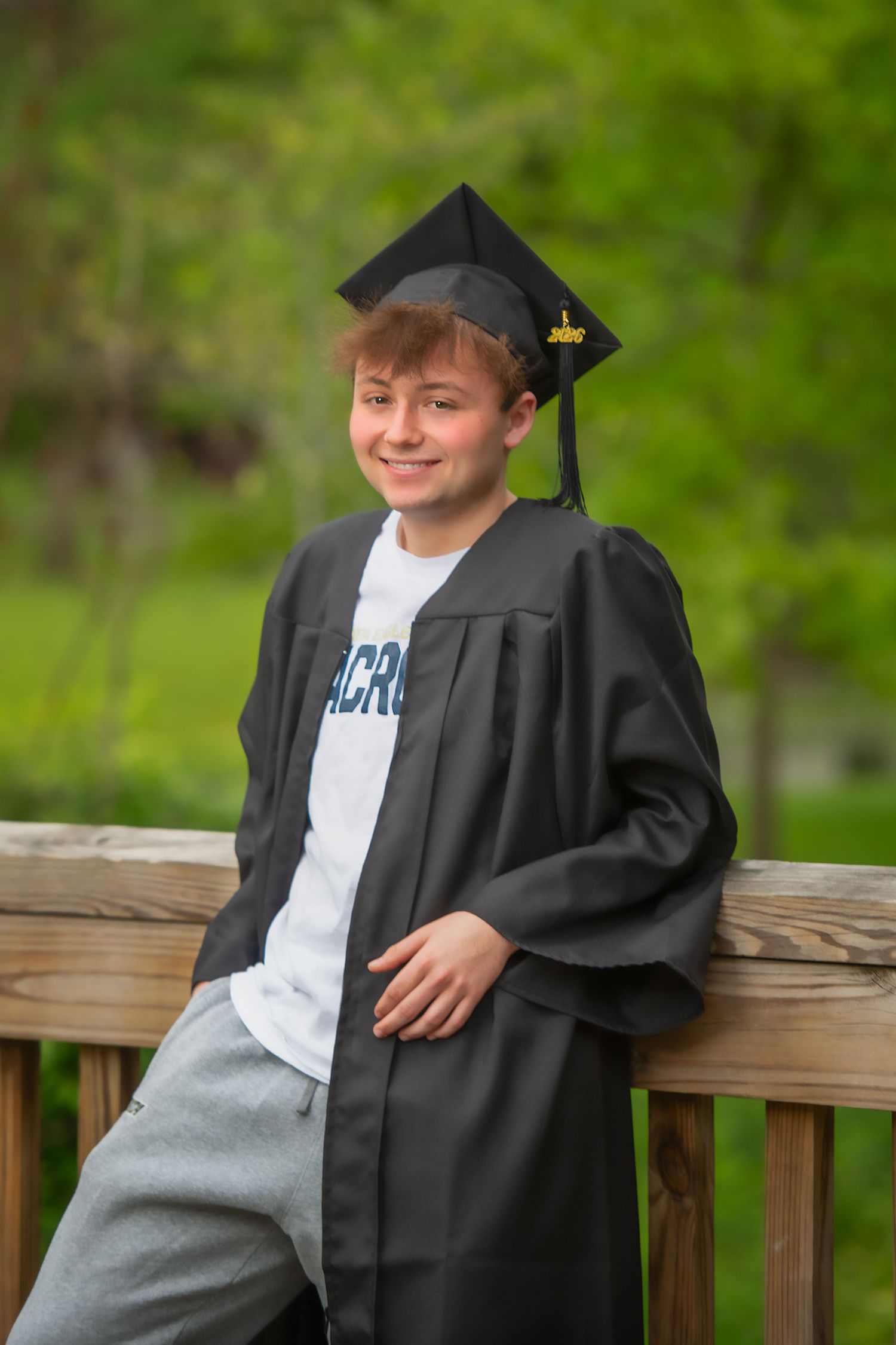 Smiling young graduate in black cap and gown leaning on wooden railing outdoors with green trees background.