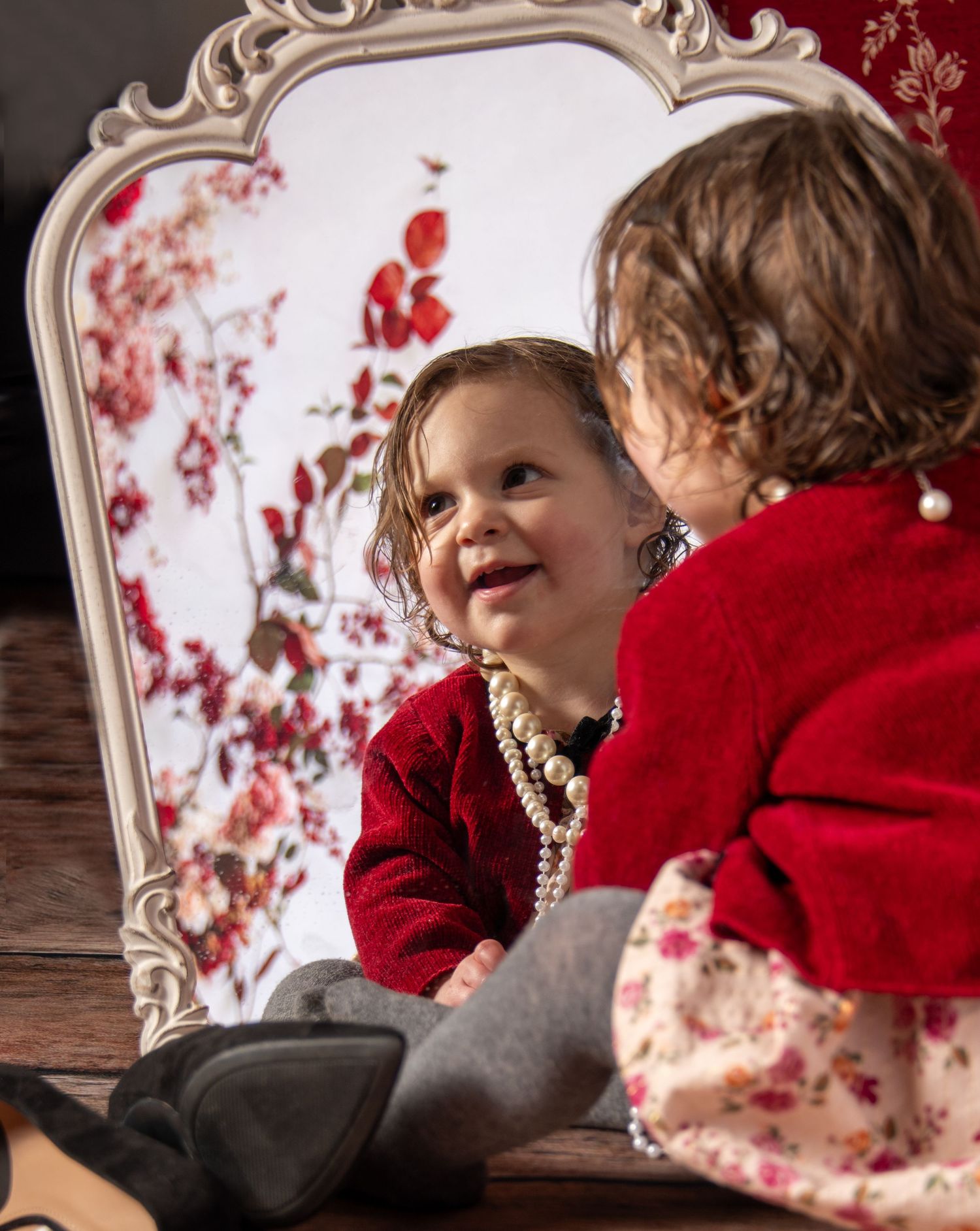 A child in a red sweater sits near a vintage mirror with floral designs and pearl necklaces.