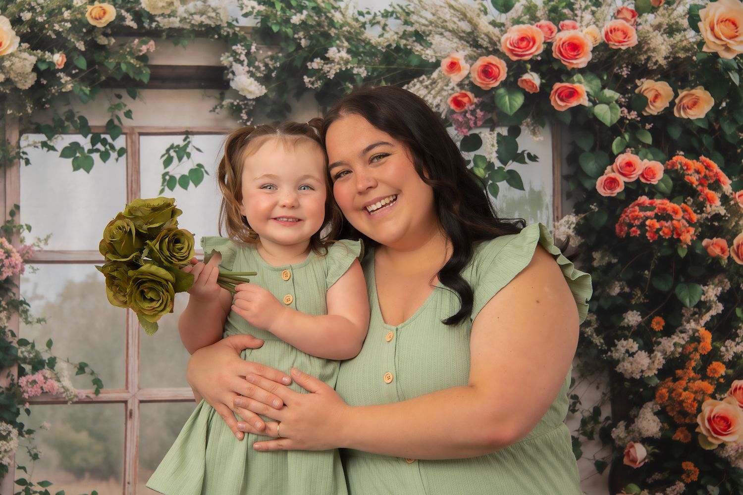 A woman and young girl in matching green dresses pose together in front of a floral rose arch backdrop.