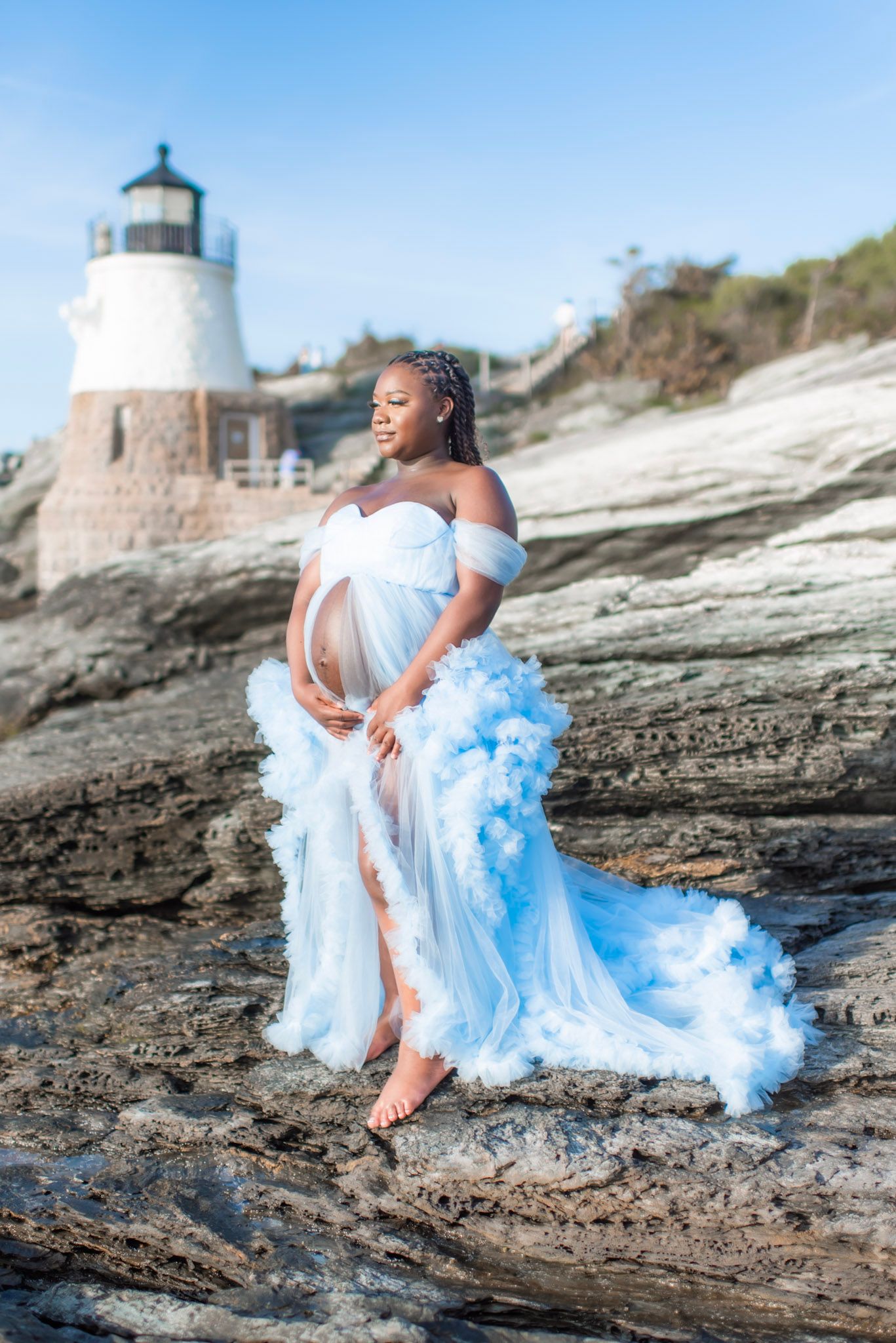 A flowing white and blue gown with dramatic train photographed against a lighthouse backdrop on rocky coastal terrain.