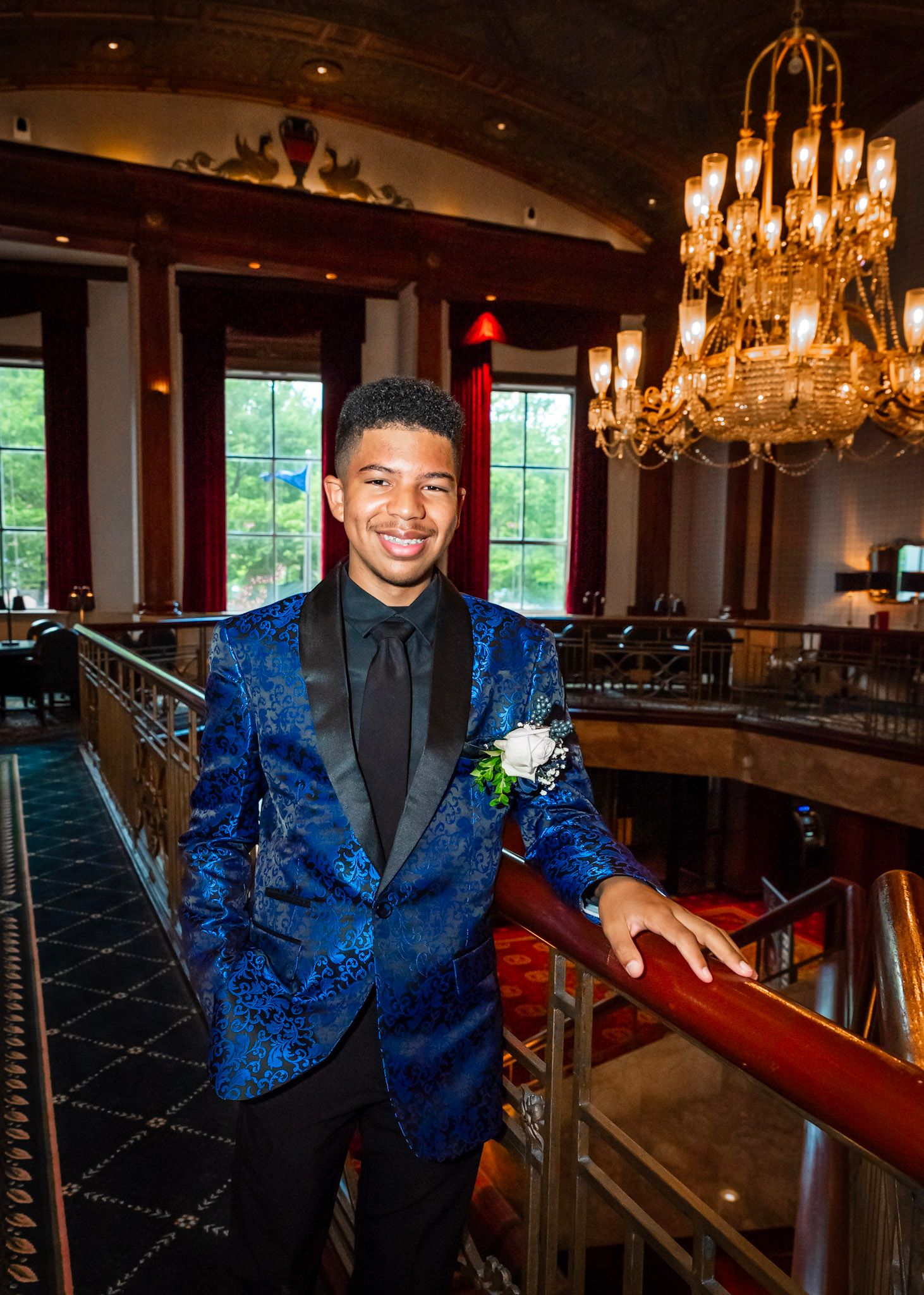 Person in blue patterned suit jacket posing in elegant venue with chandeliers and windows.