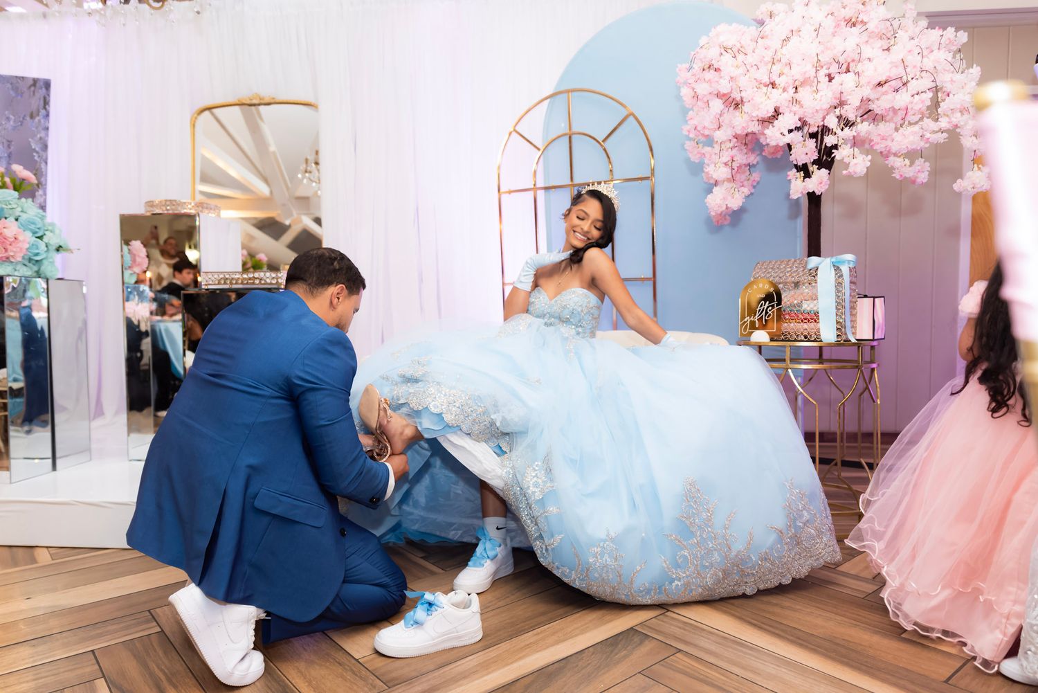 Indoor photoshoot with pink cherry blossoms and light blue princess gown. Father changing the gir's shoes at a sweet 16