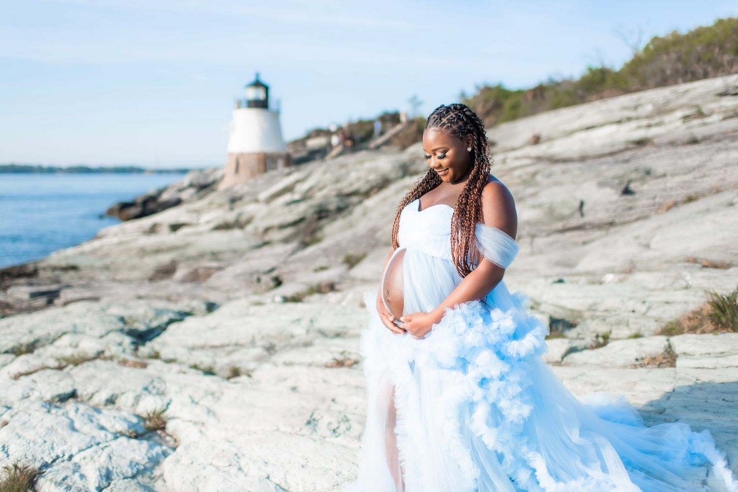 Coastal maternity photoshoot with flowing blue dress against rocky shoreline and lighthouse.