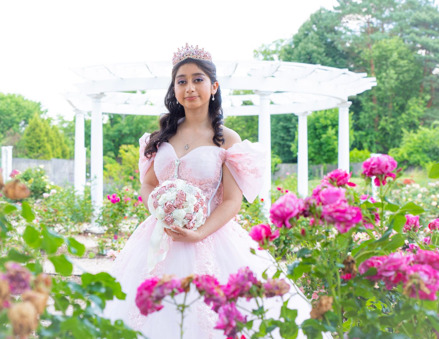Quinceañera photoshoot in Garden setting with white pergola and vibrant pink roses in full bloom.