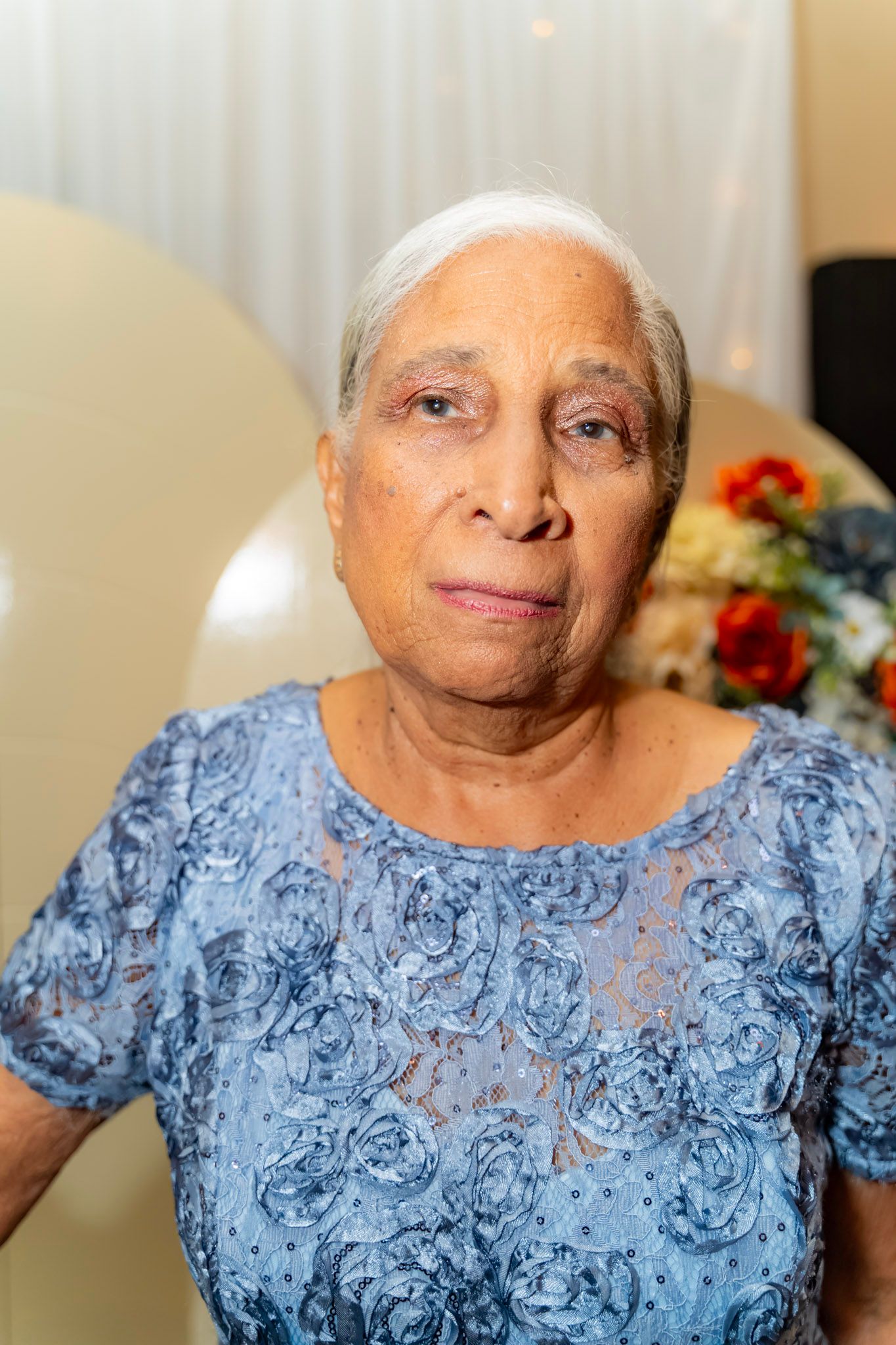 An elderly person wearing a light blue floral lace top poses for a portrait with a gentle expression.