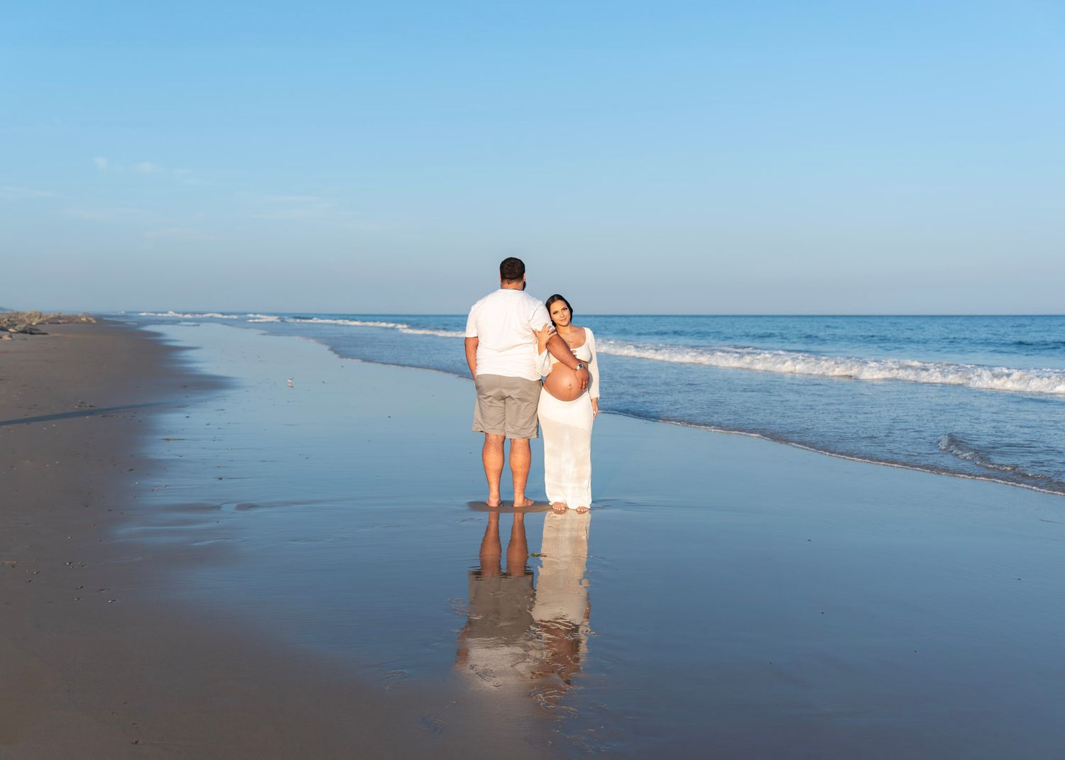 Maternity Photoshoot featuring Peaceful beach scene with gentle waves rolling onto shore during golden hour. Mom sweetly holding dad's arm.