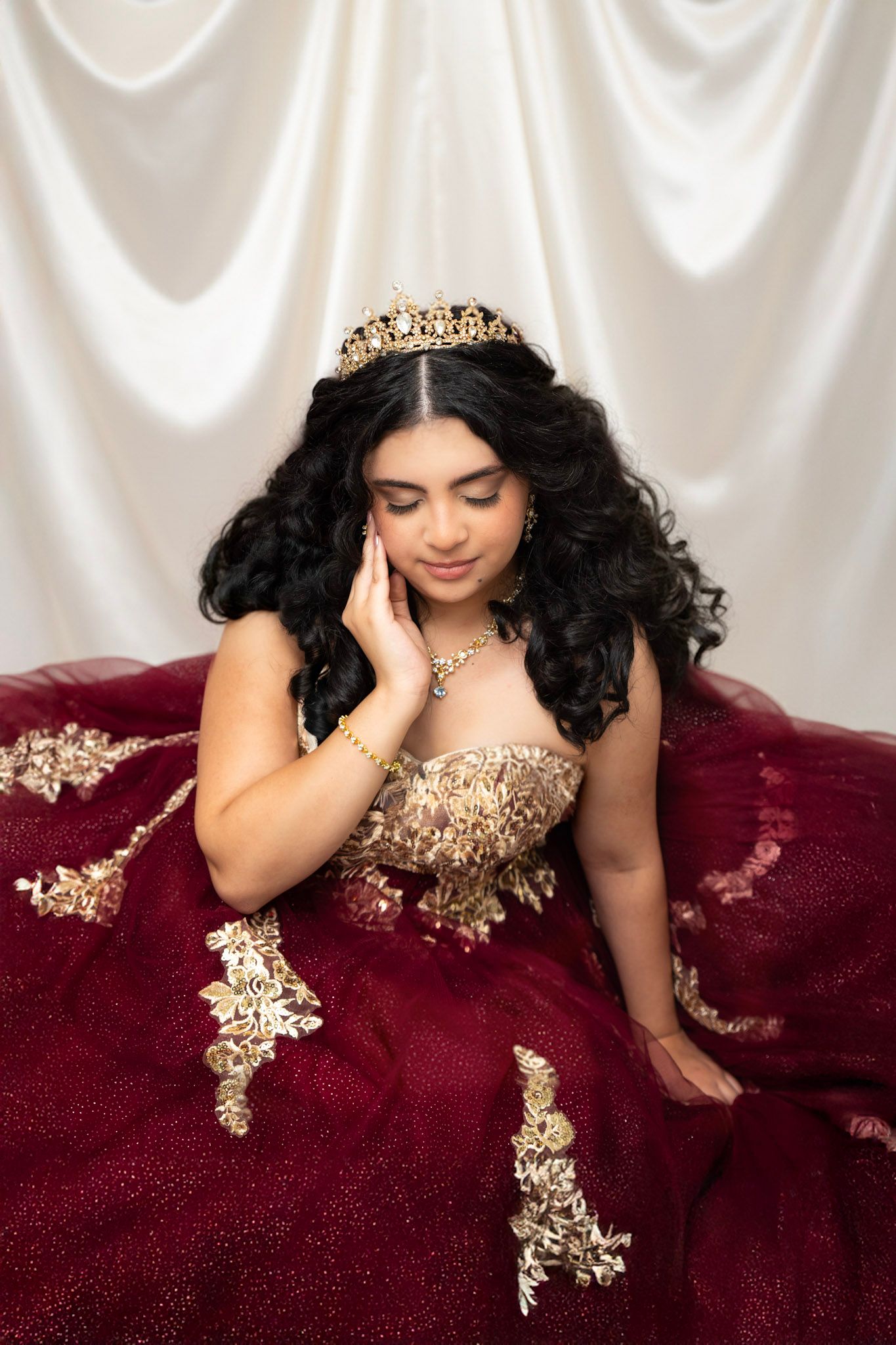 Elegant portrait on white satin backdrop wearing gold embellished burgundy gown.