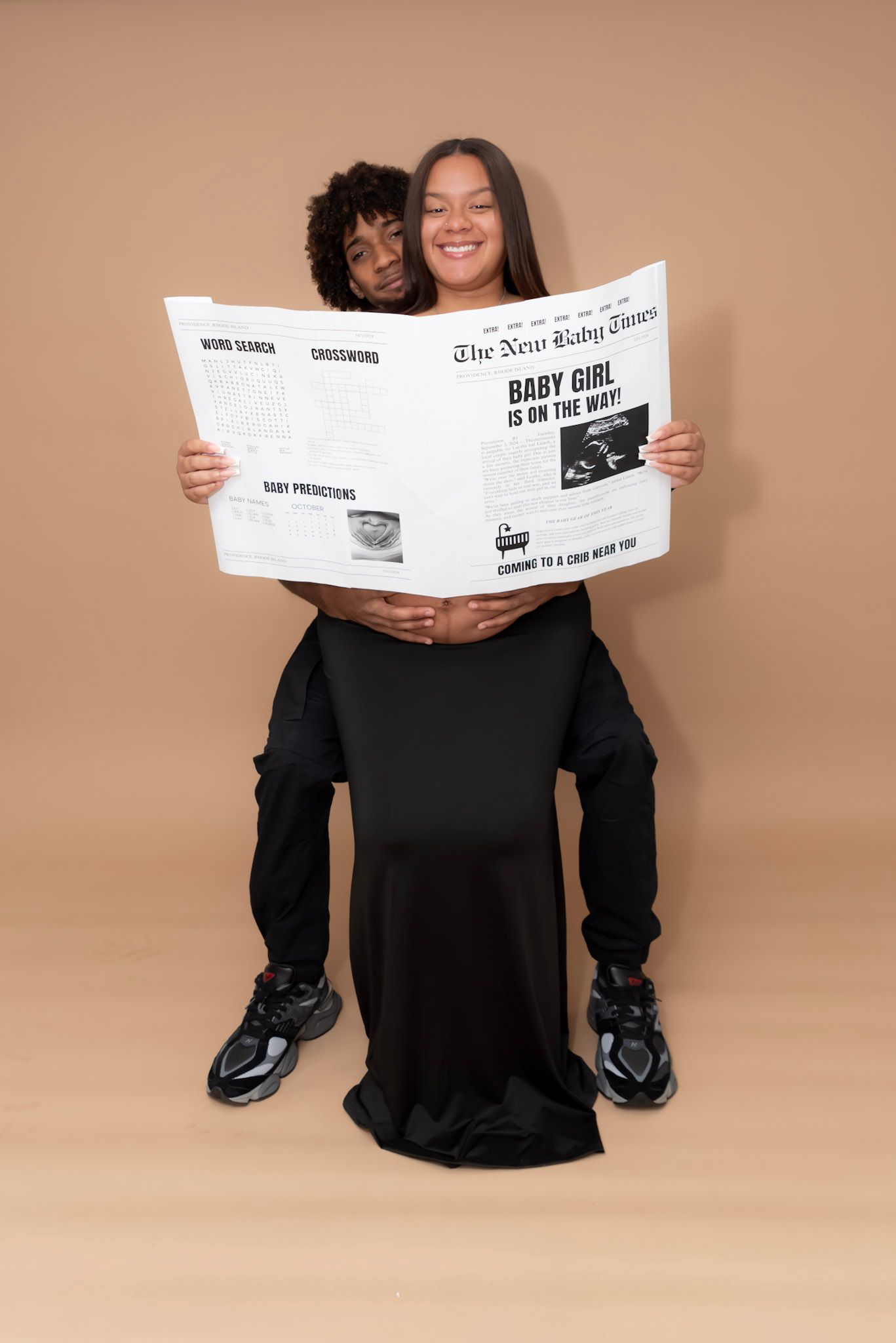 Creative maternity photo with newspaper and black dress against beige backdrop.