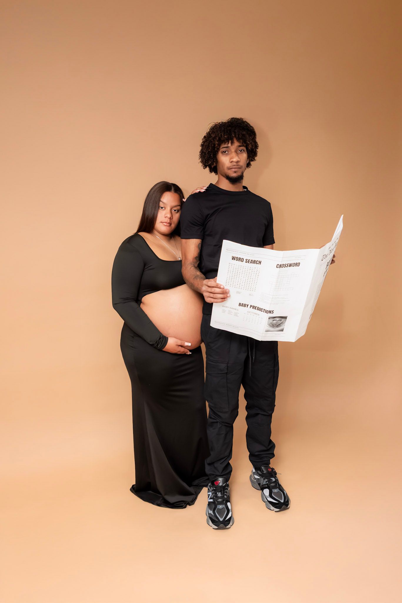 A couple poses against a beige backdrop, with one reading a newspaper while the other shows off a pregnant belly.