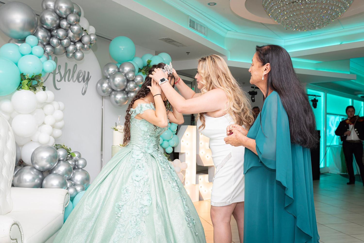 A celebration scene with teal and silver balloon decorations and a sparkling chandelier in an elegant venue.