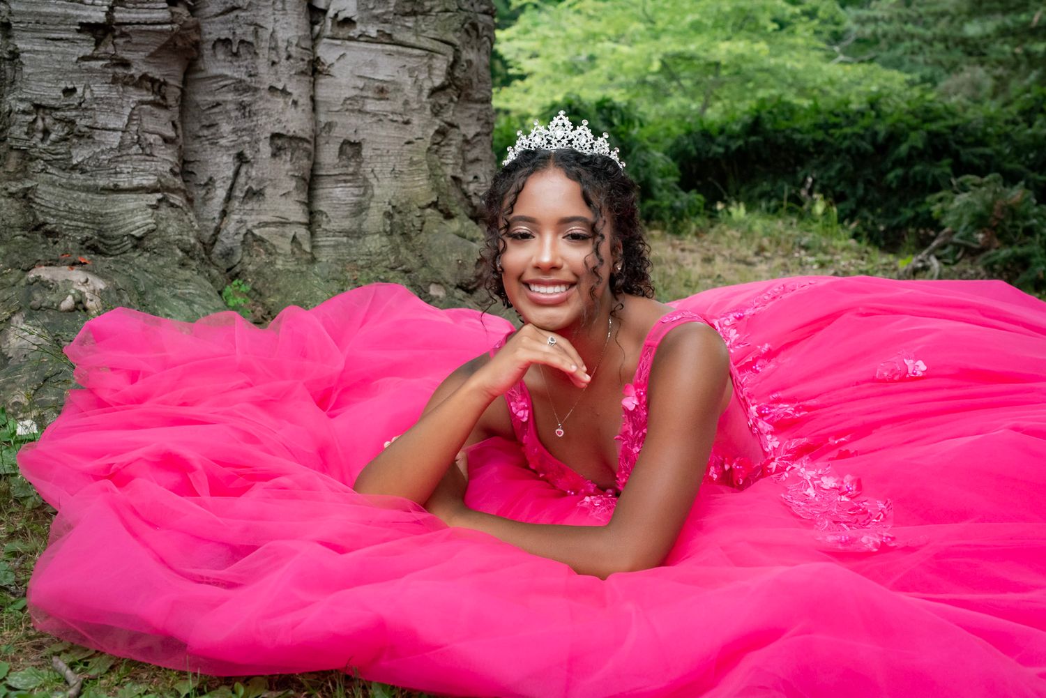 A person in a bright pink ball gown and tiara poses gracefully against a stone wall backdrop in a garden setting.