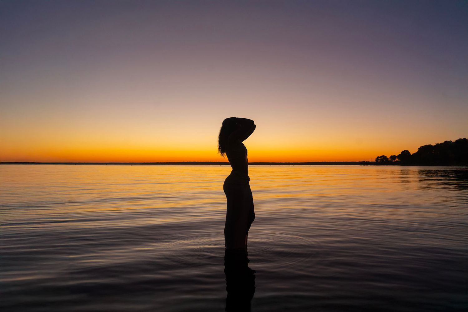 Silhouetted figure stands on calm water against vibrant orange and purple sunset sky over lake horizon.