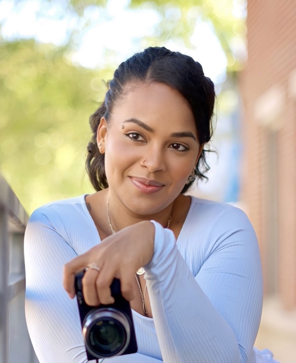 Professional photographer in casual blue top holding camera in outdoor setting.